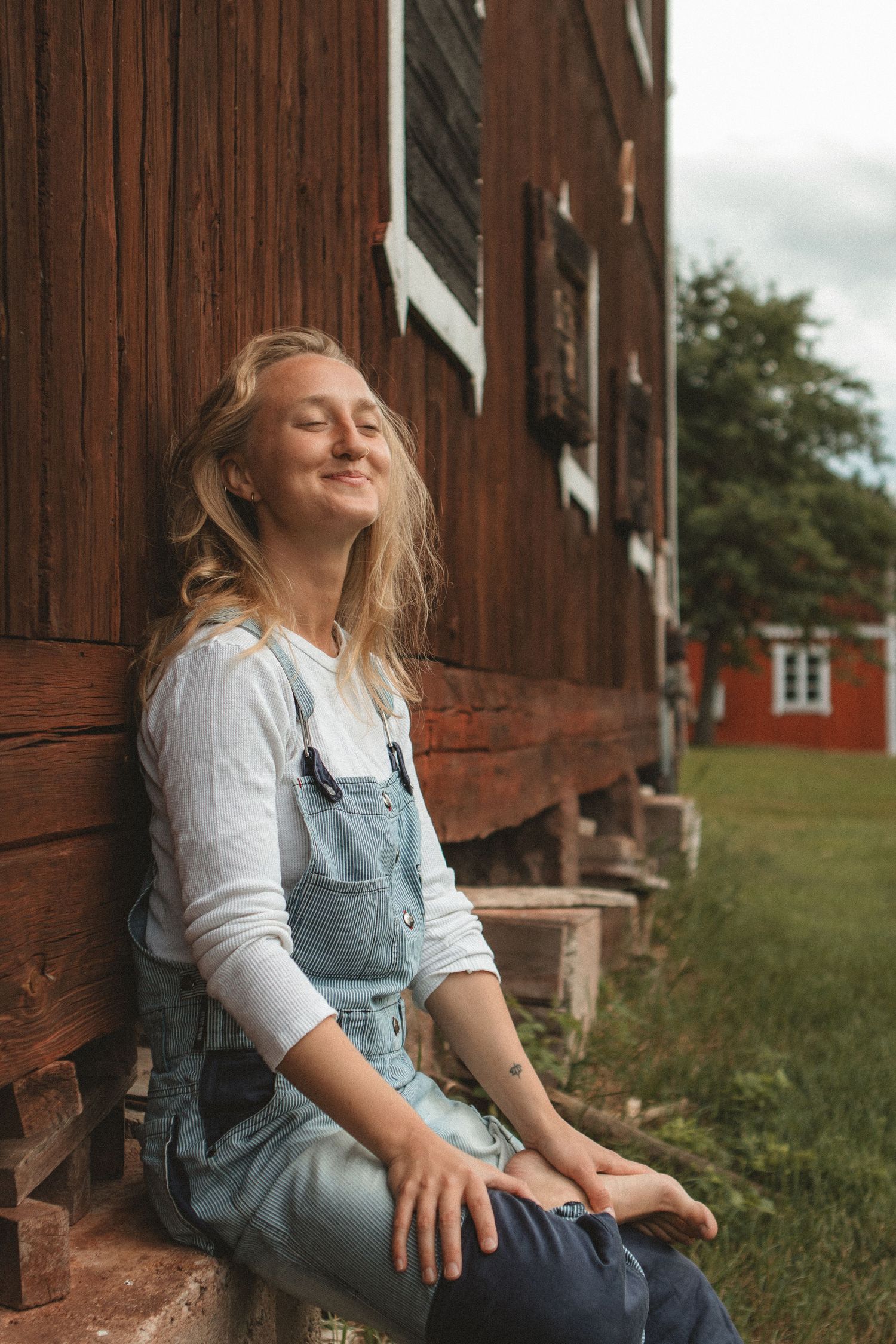 Person in denim overalls and white sweater rests on steps of rustic barn building.