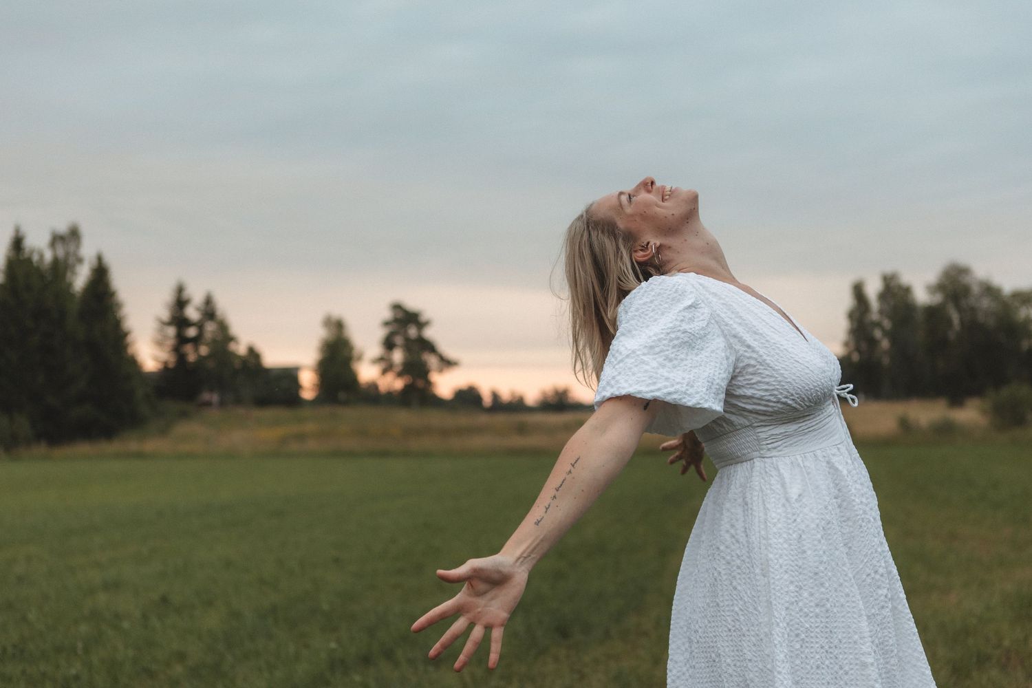 A person in a white dress stands in a grassy field at sunset with arms outstretched.