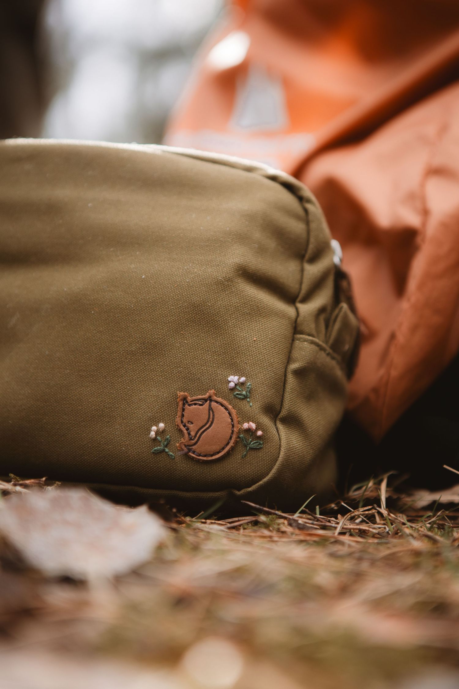 Olive green Fjallraven bag with embroidered fox logo resting on forest ground beside an orange backpack.