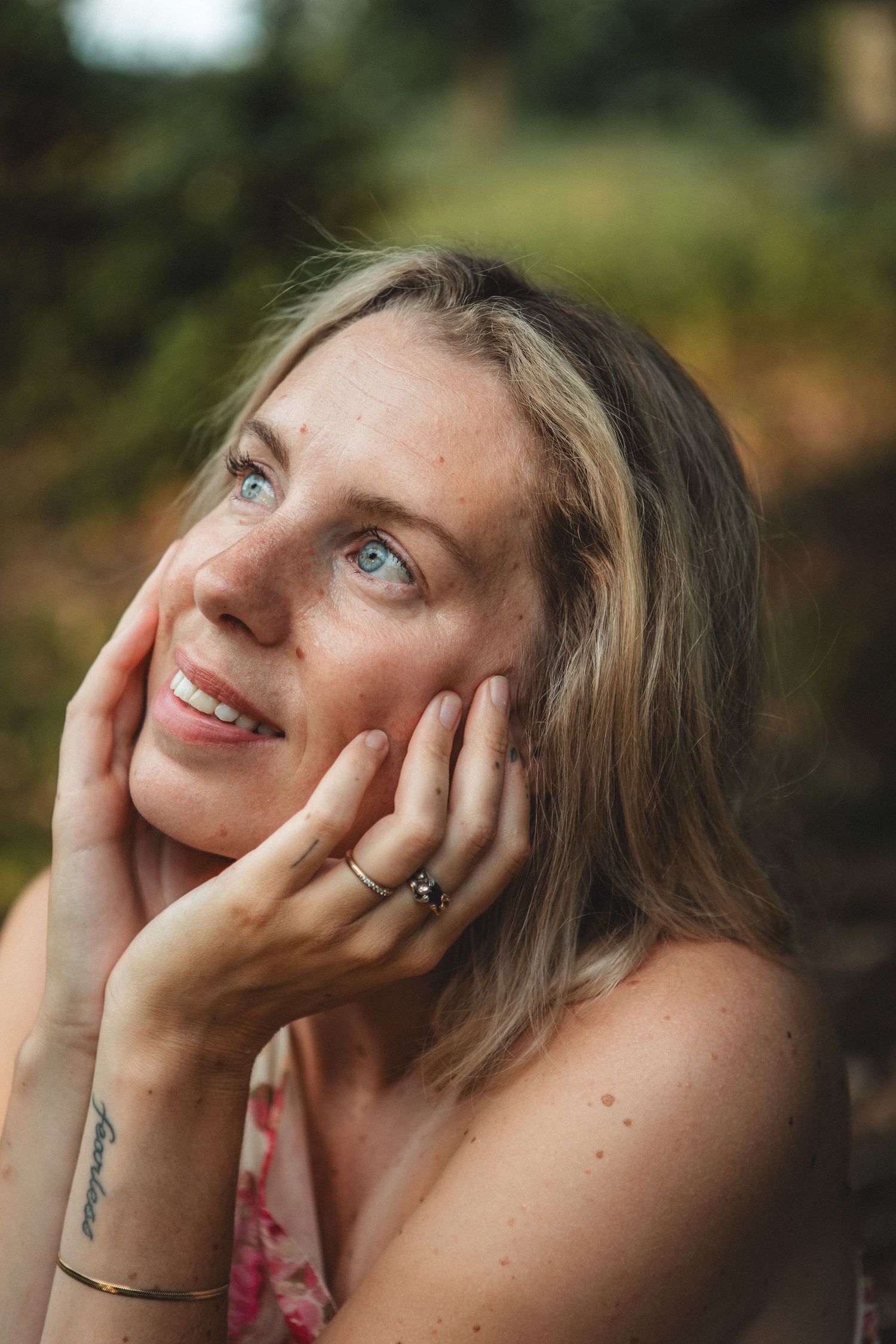 A person with blonde hair smiles while touching their face, wearing a floral dress outdoors.