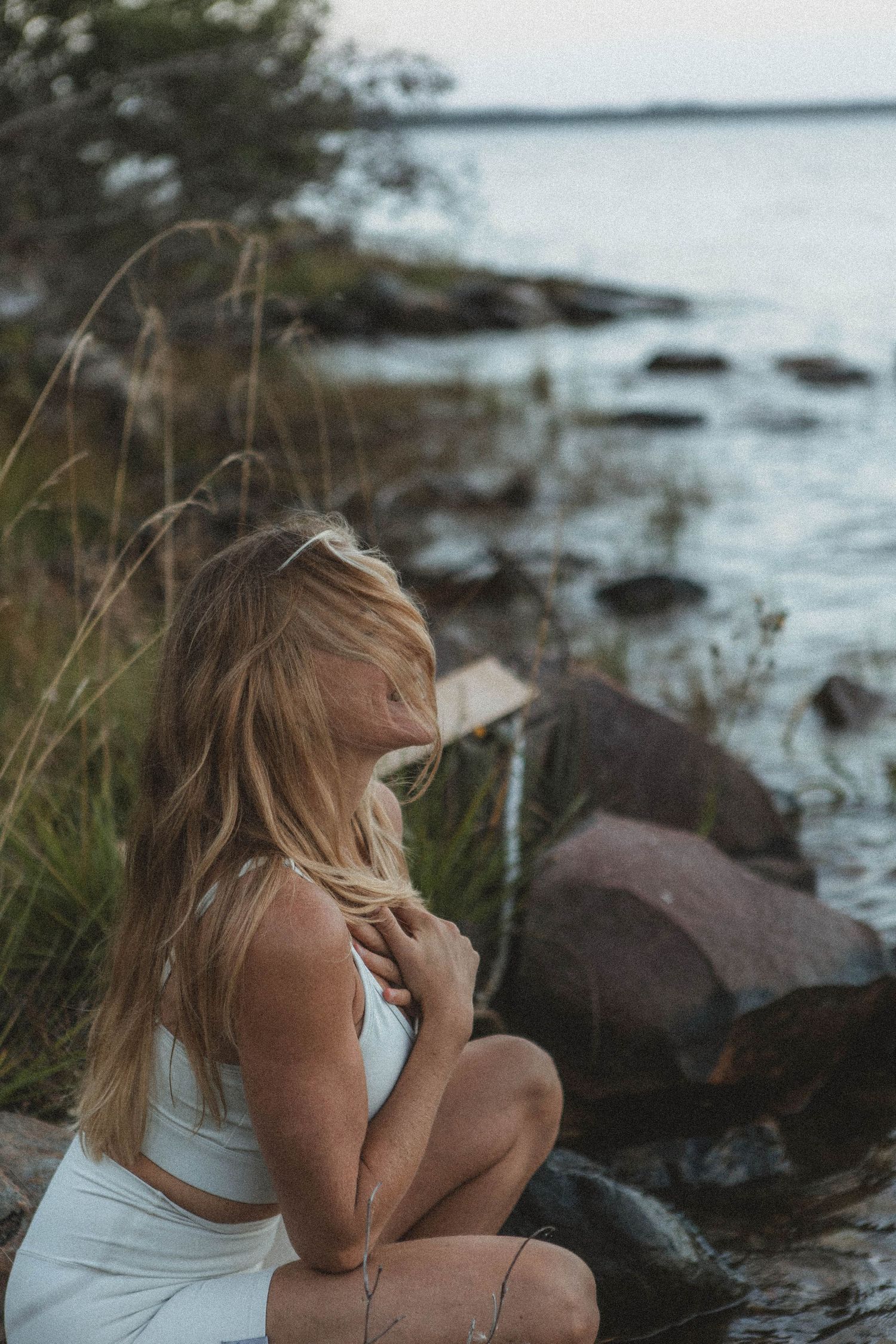 Person in white athletic wear sits thoughtfully by ocean rocks at sunset.