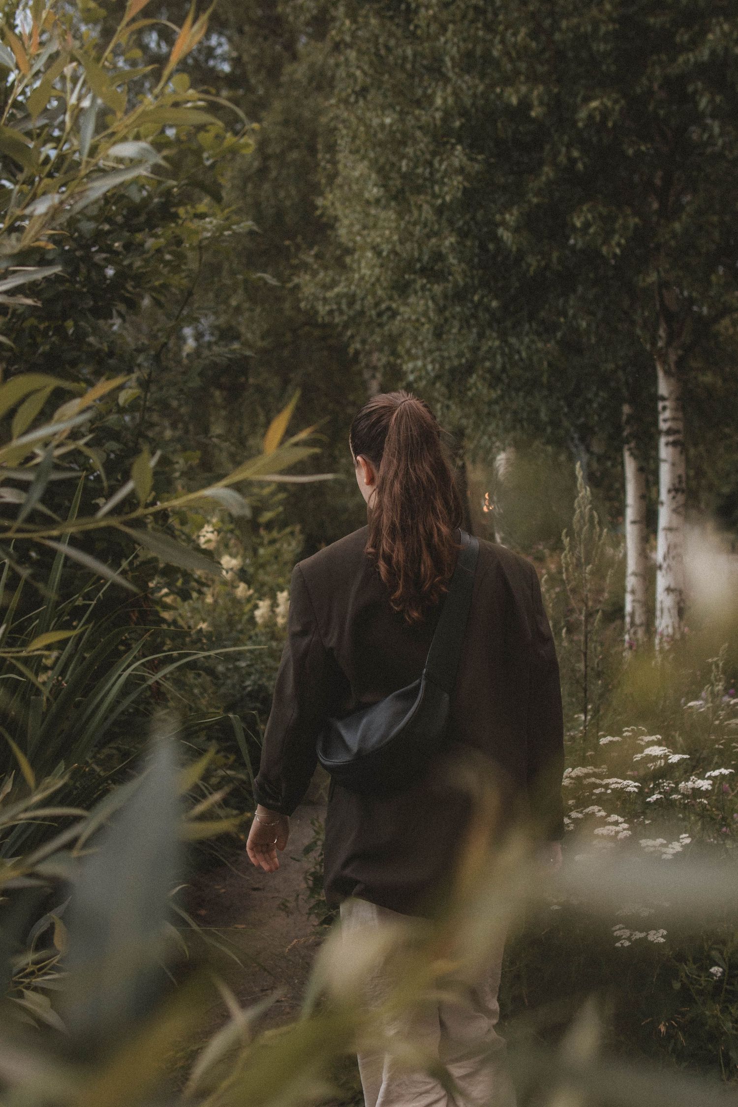 Someone wearing a black sweater walks along a misty forest path bordered by birch trees and tall grasses.