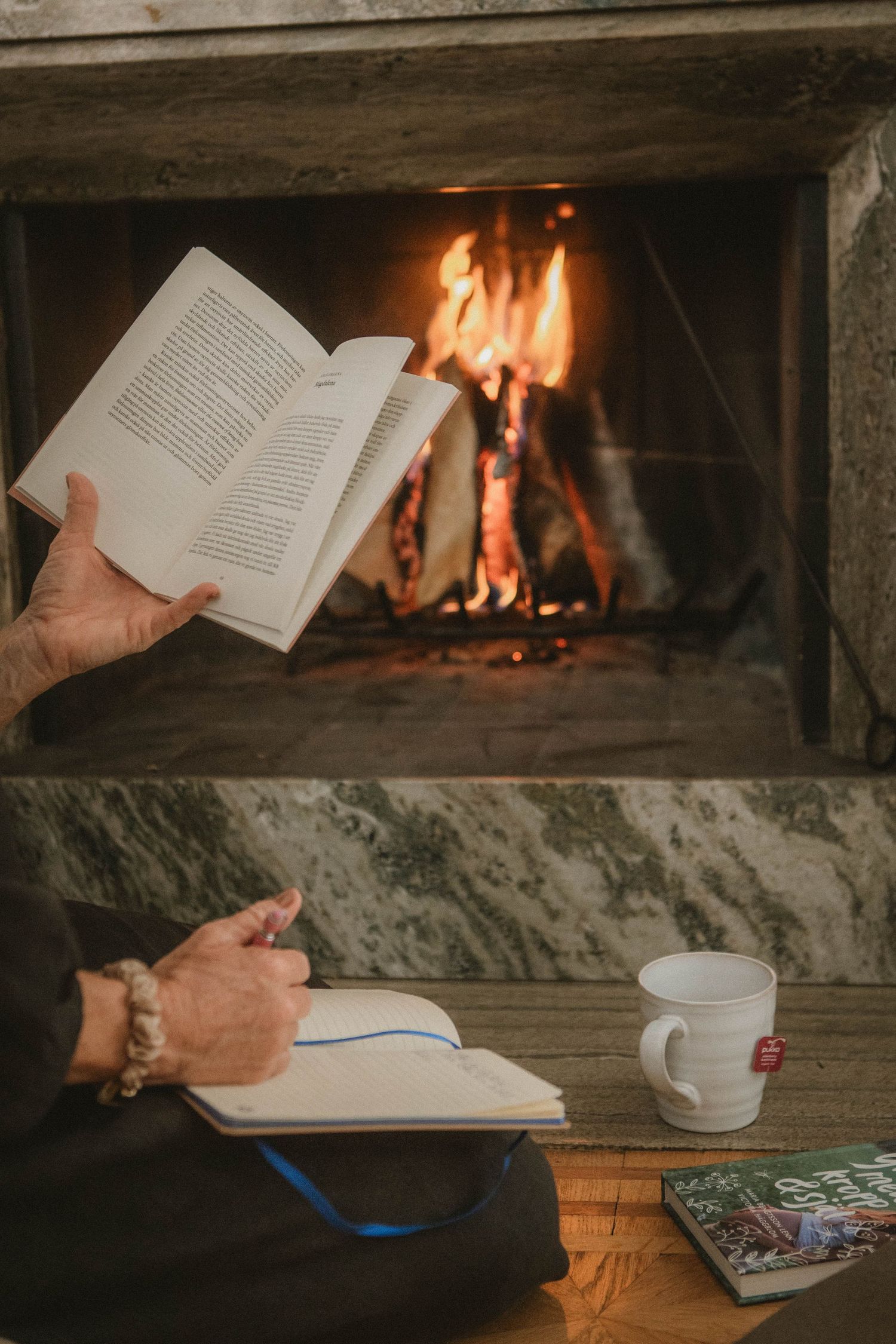 Open book held near cozy fireplace with coffee cup and journal on wooden surface.