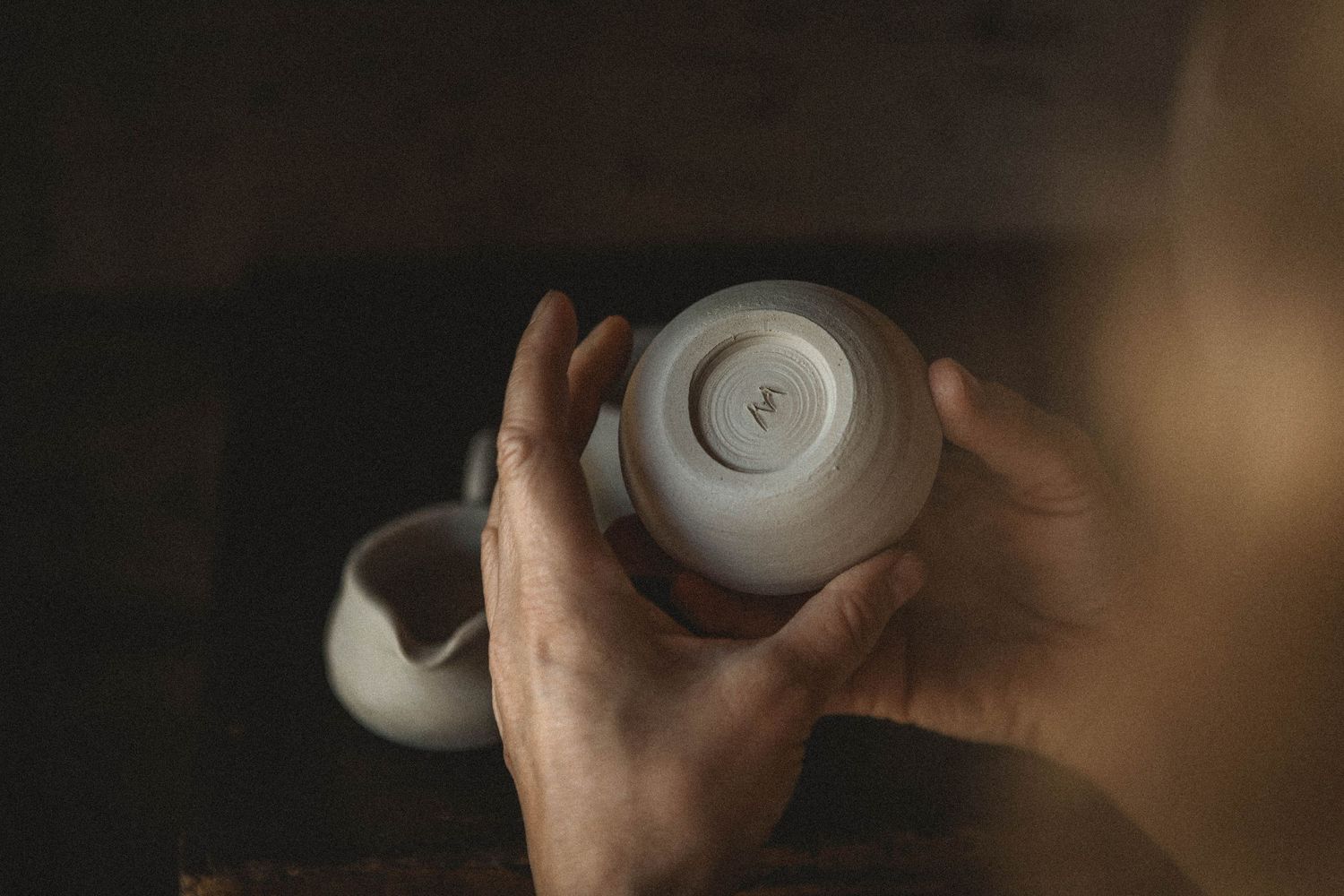 Hands hold a white ceramic vessel in moody lighting against a dark background.
