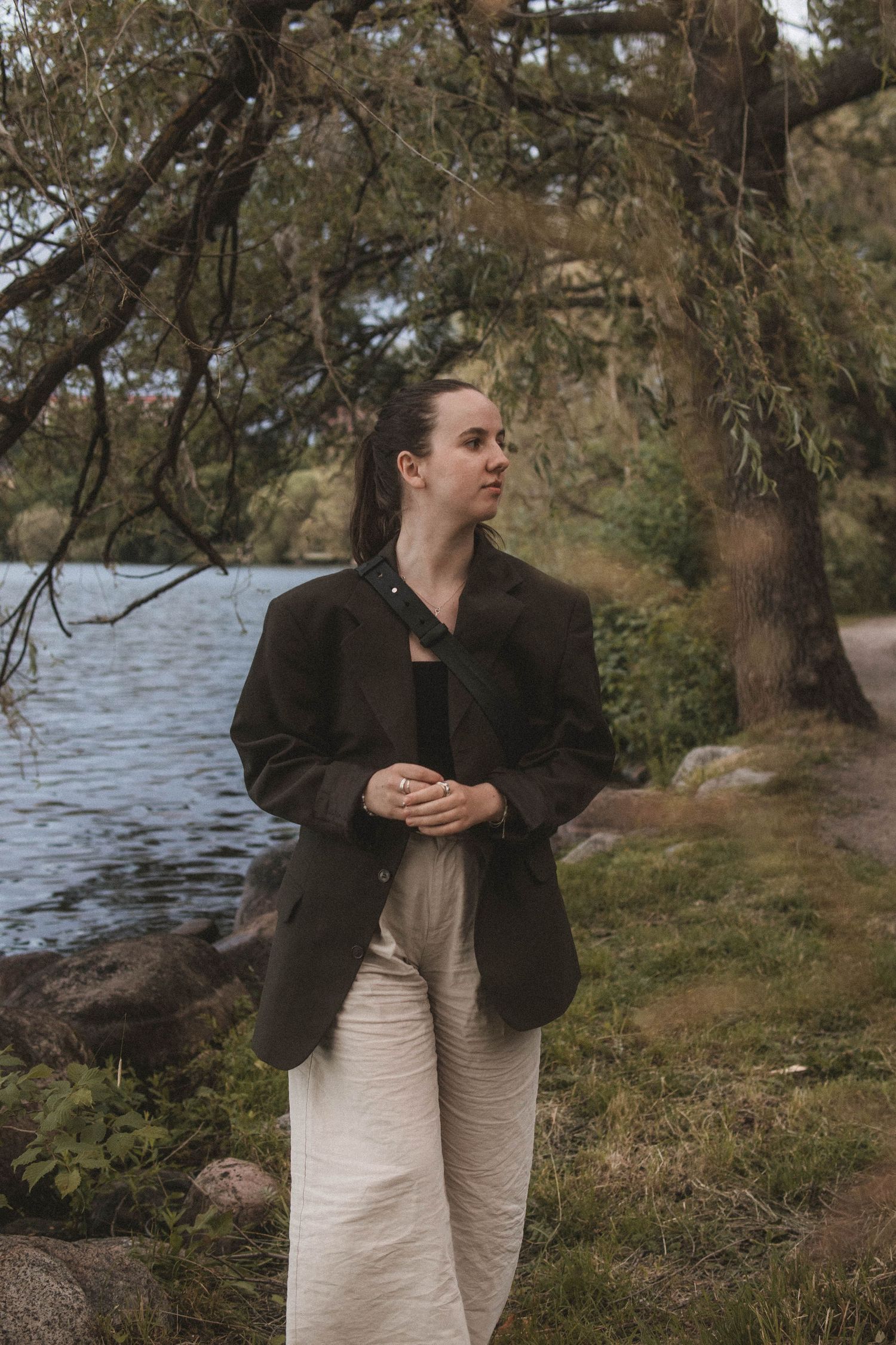 Person in dark blazer and light pants stands thoughtfully by a lakeside with trees in the background.