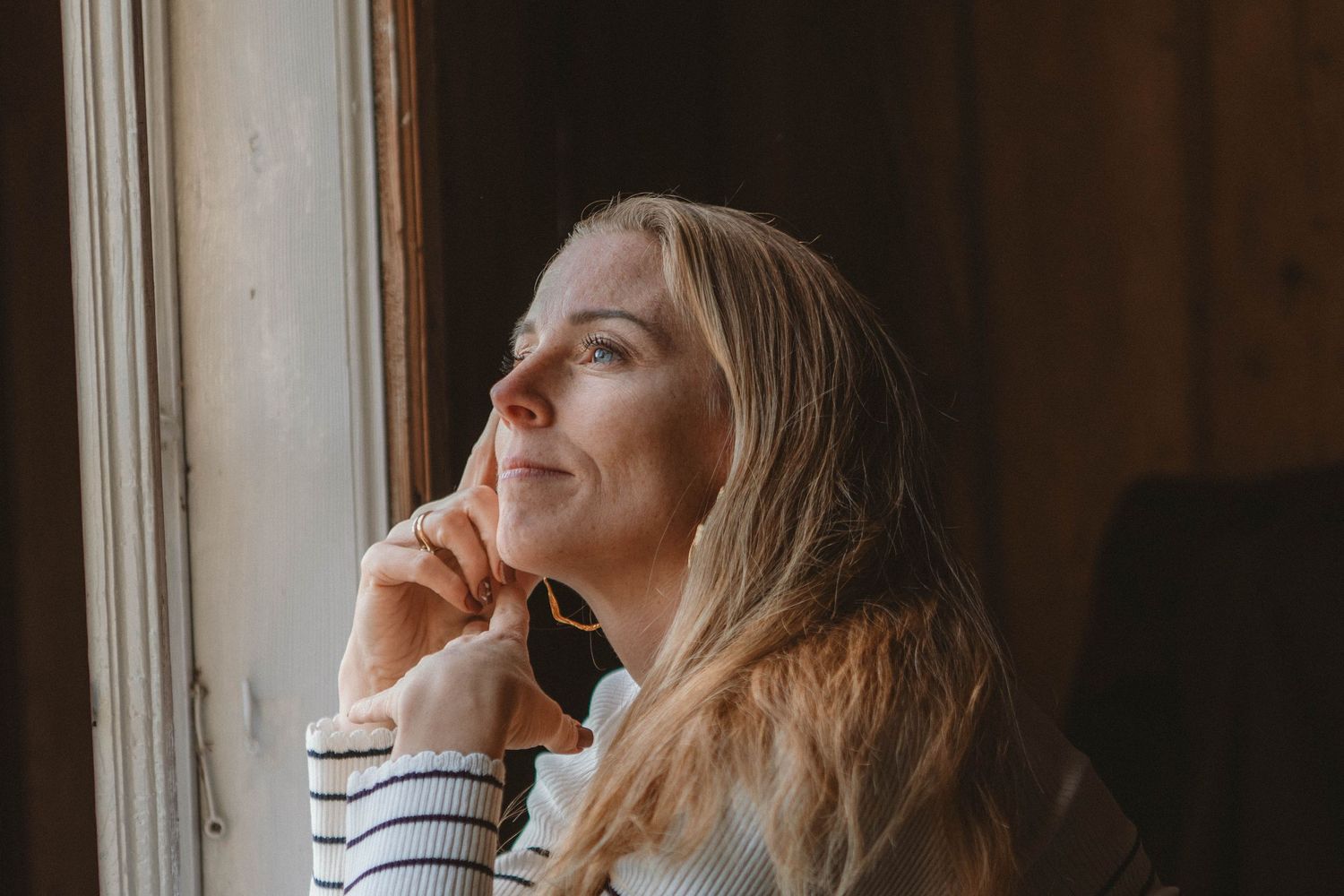 Person in striped sweater gazes thoughtfully out window in warm natural light.