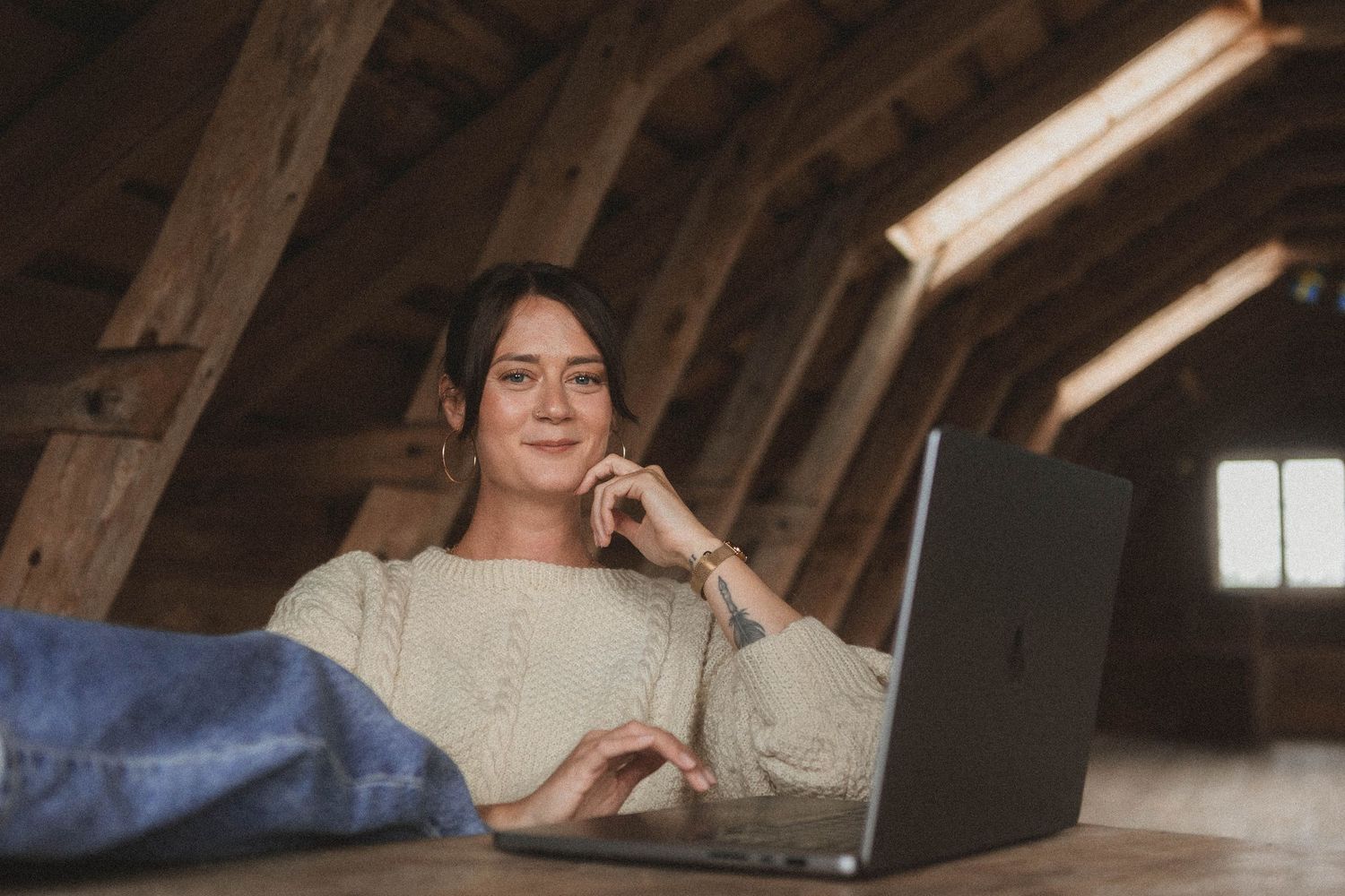 Person working and smiling at laptop computer in rustic attic space with natural lighting coming through window.