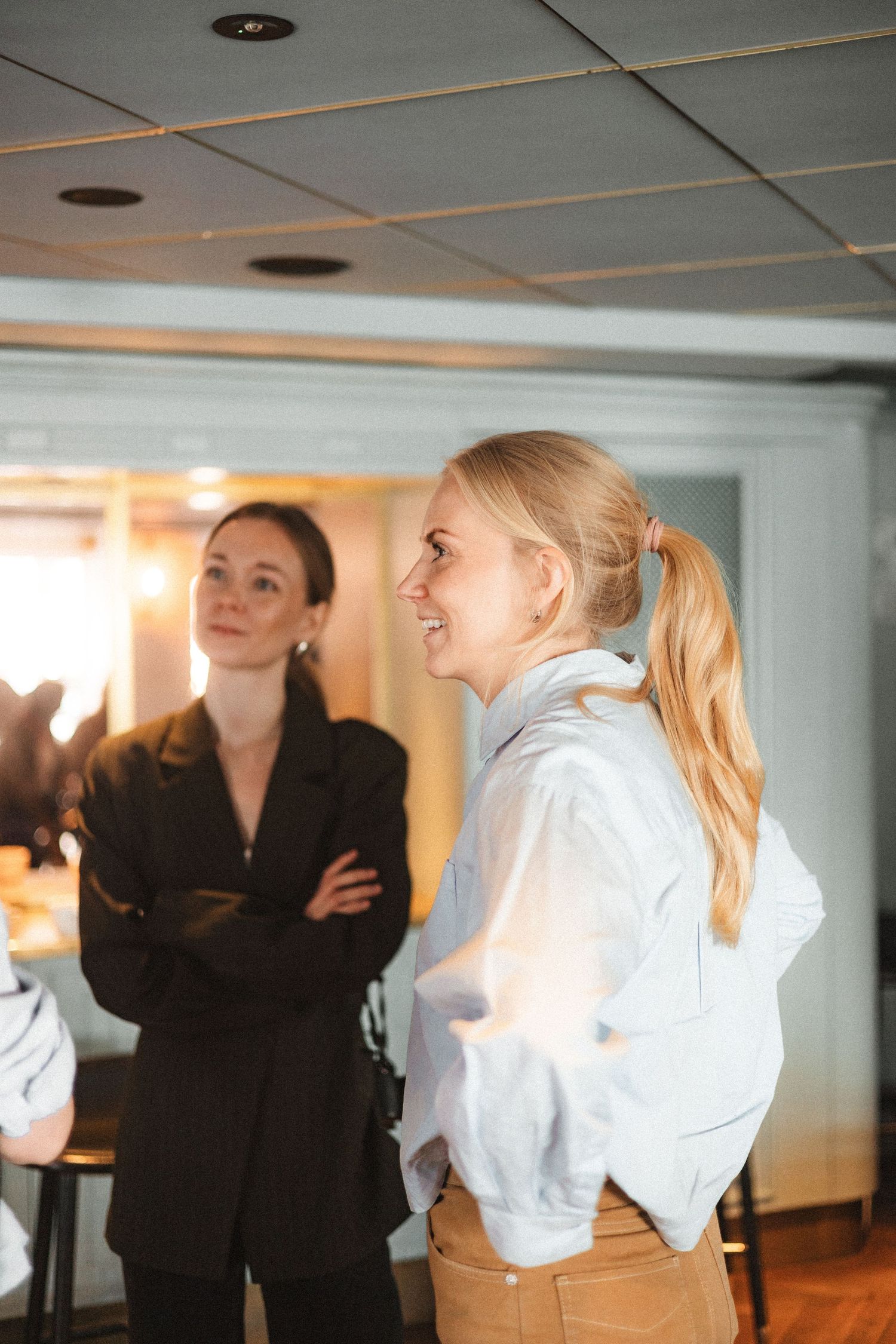 Two individuals engage in conversation while standing in an office hallway with fluorescent lighting.