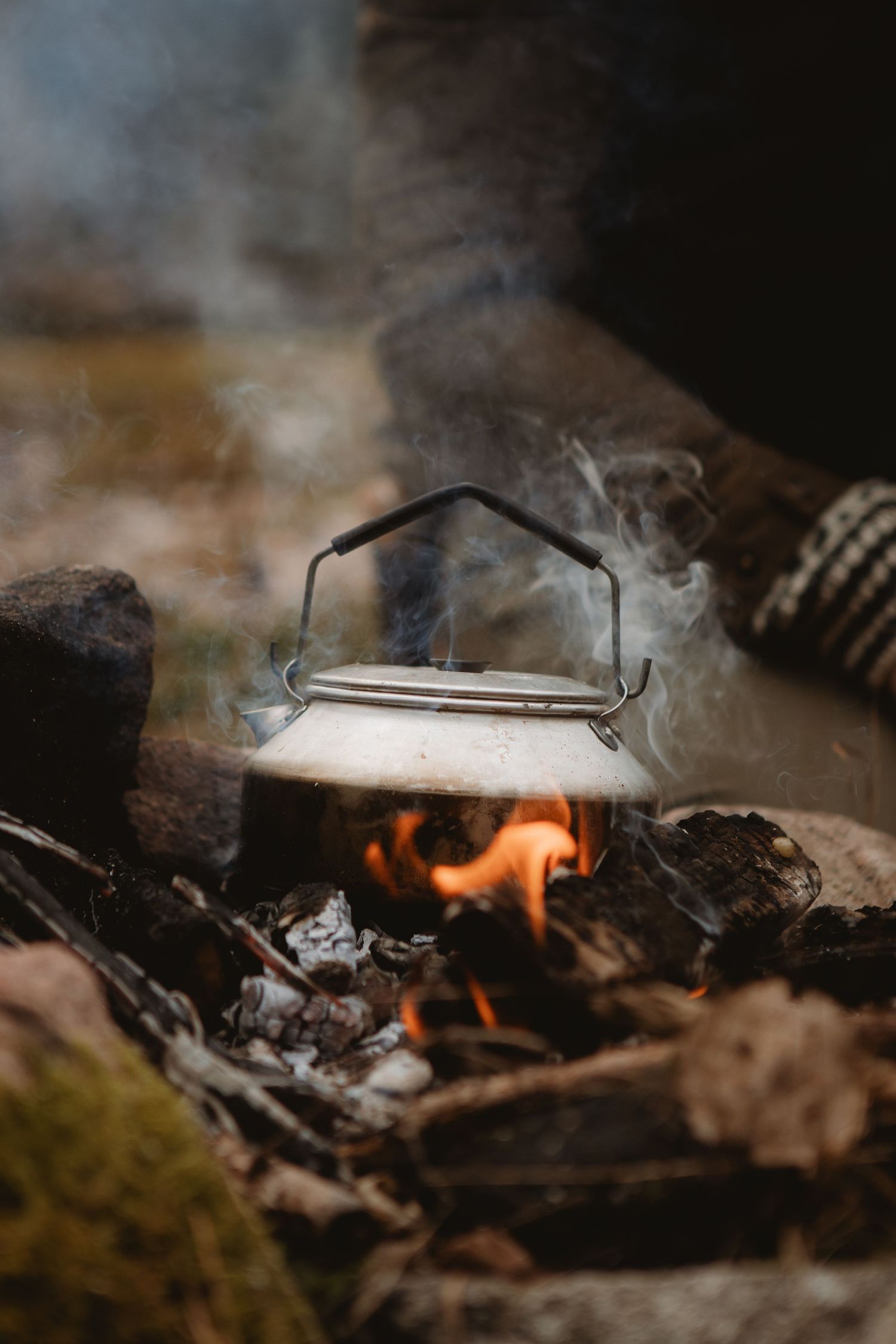 A vintage metal kettle boiling over an open campfire surrounded by rocks outdoors.