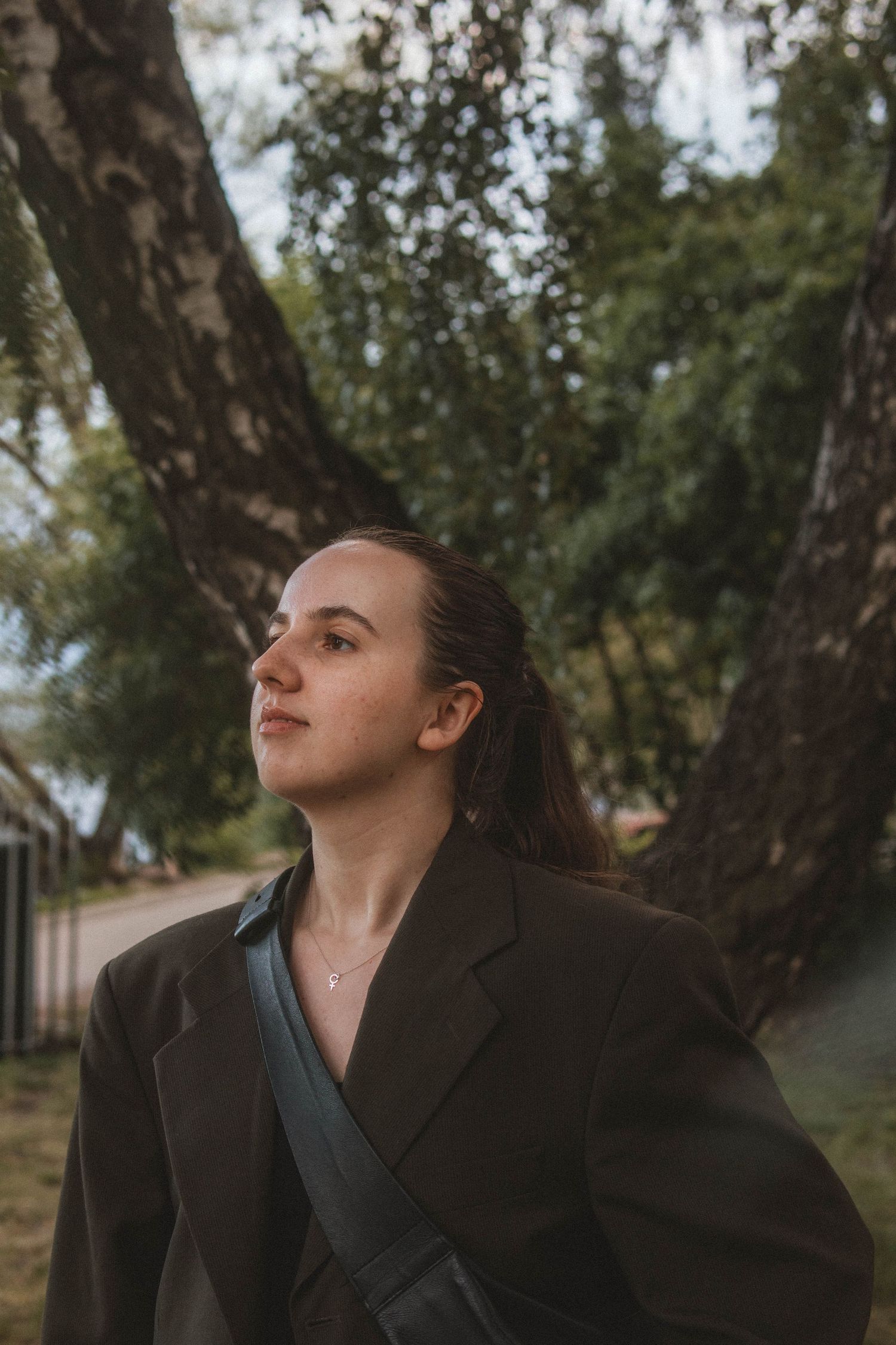 Portrait of a person in a brown blazer looking up thoughtfully against a blurred natural background with trees.