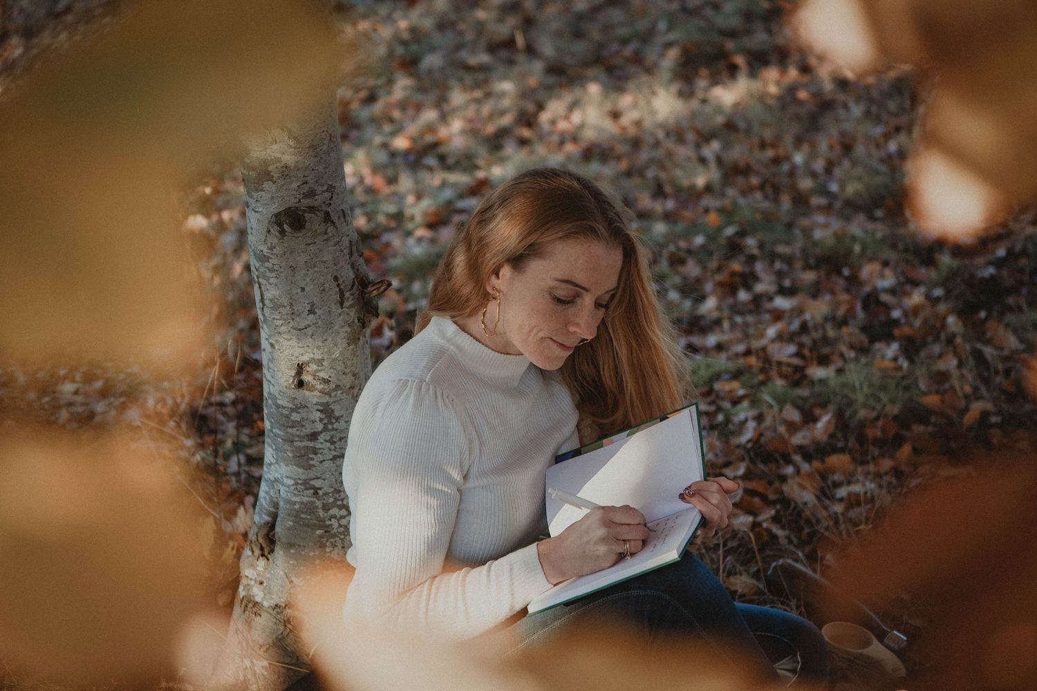 Someone in a white sweater writes in a journal while sitting against a birch tree trunk on an autumn day.