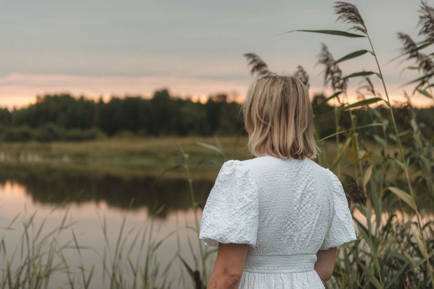 Someone in a white dress stands near reeds by a lake during sunset.
