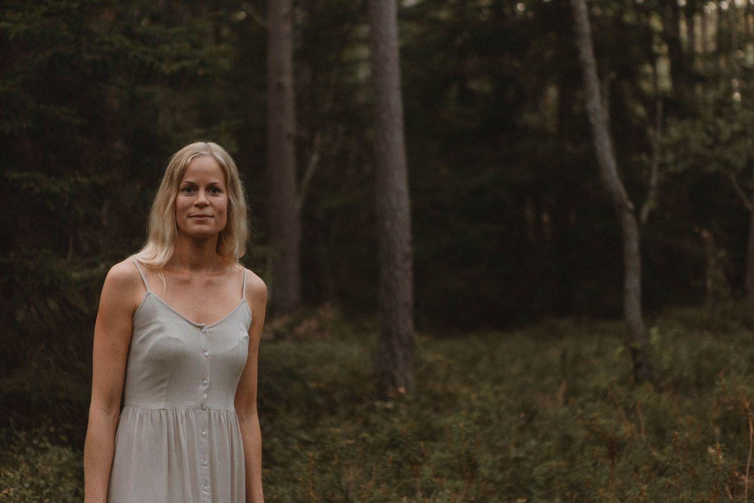 Figure in light dress stands still among tall pine trees in moody forest lighting.