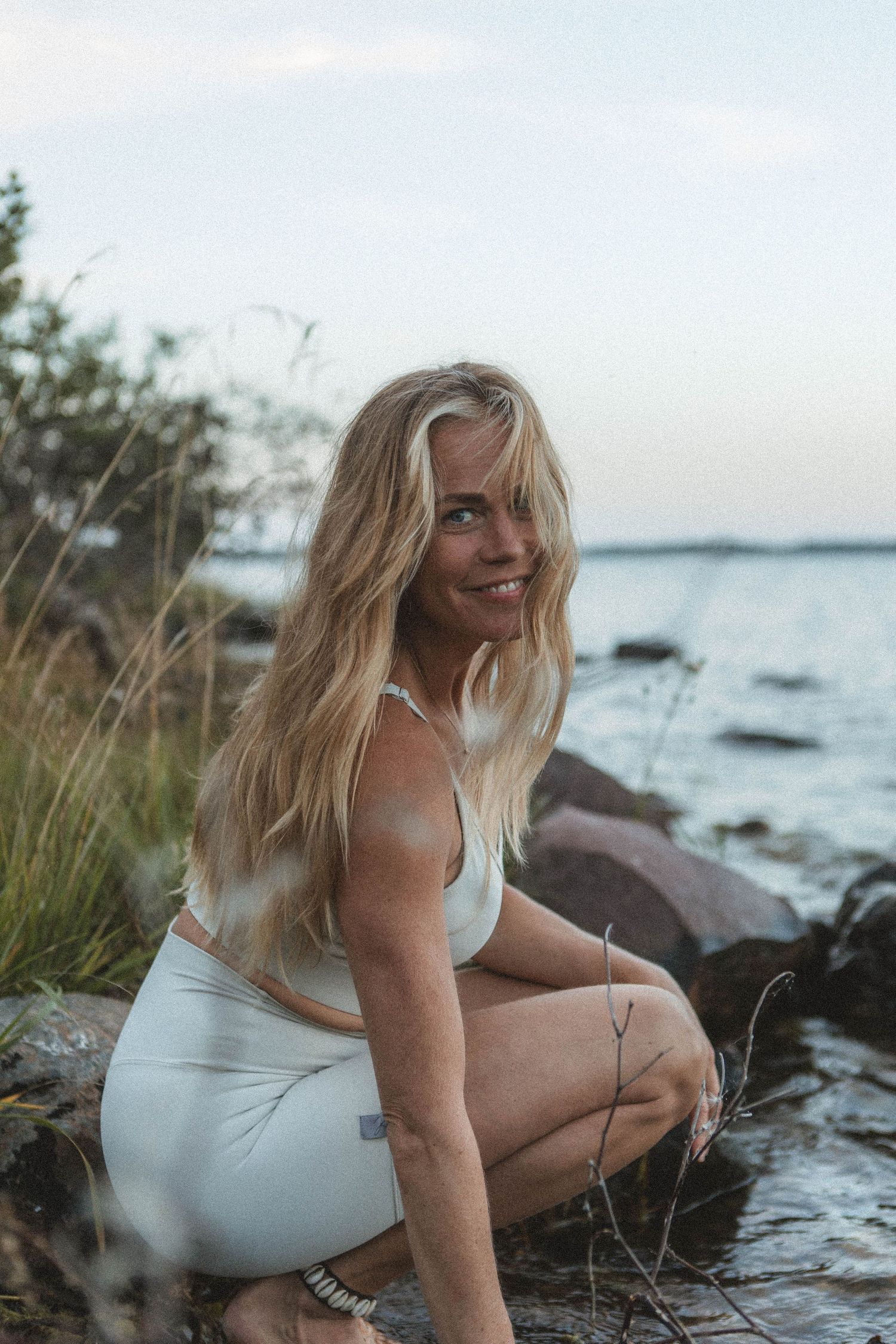 Athletic figure in white activewear poses by rocky shoreline at dusk.