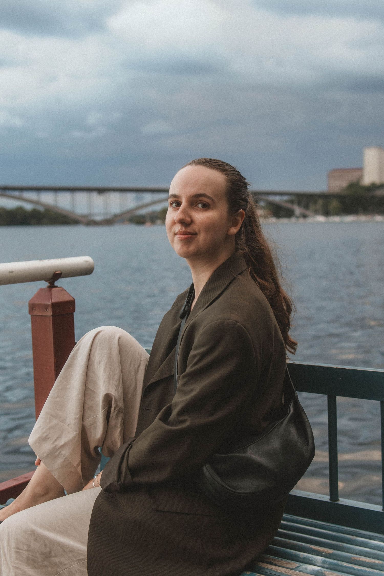 A person in an oversized blazer sits on a bench near water with a bridge visible in the background.