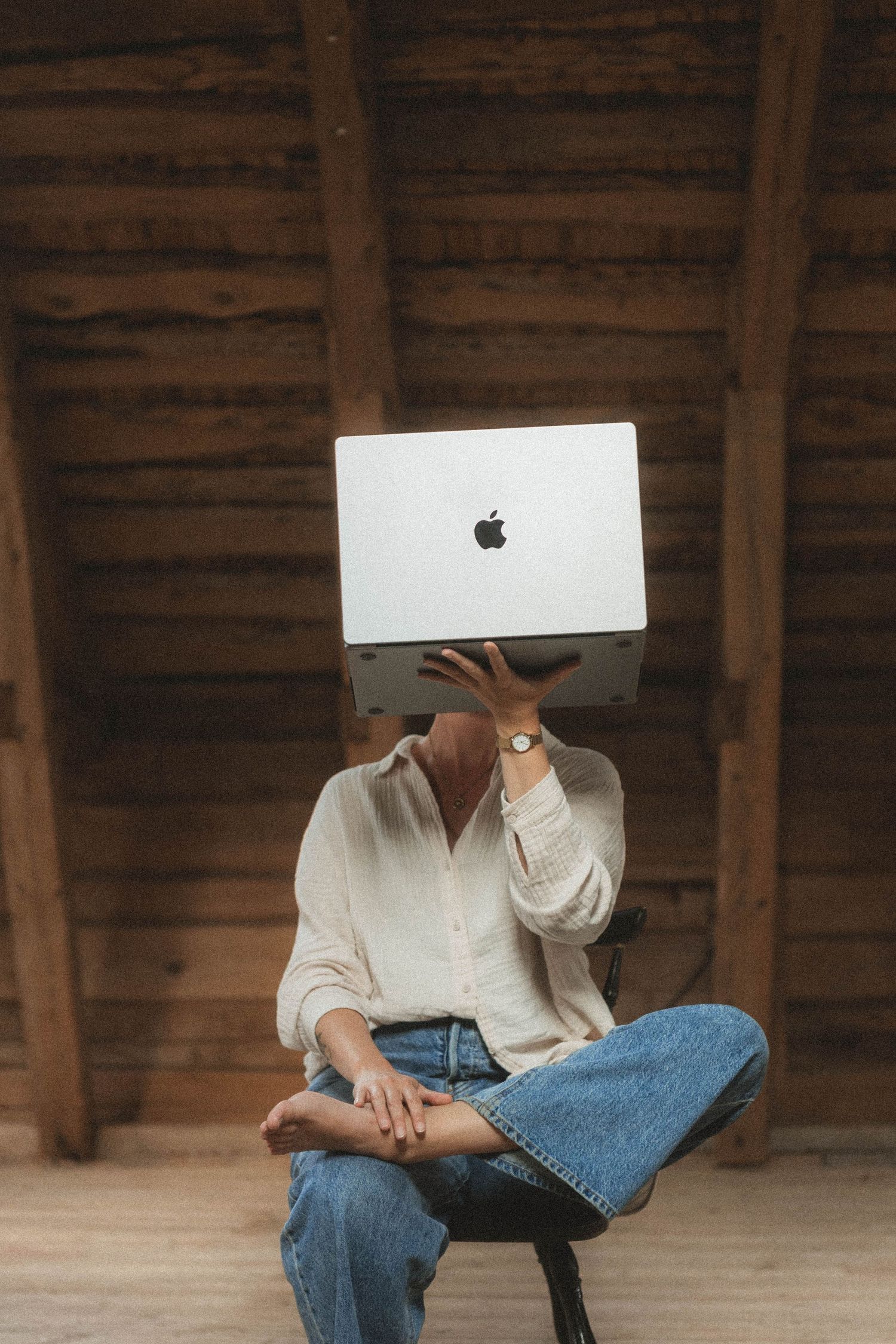 Person working on MacBook laptop while sitting in wooden chair in rustic attic interior.