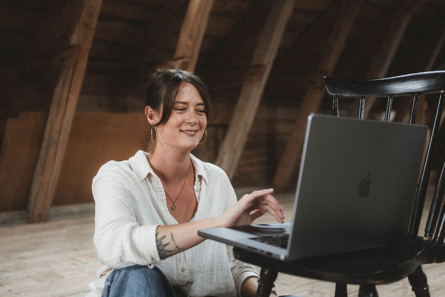 A person in a white shirt smiles while working on a laptop in a rustic wooden attic space.