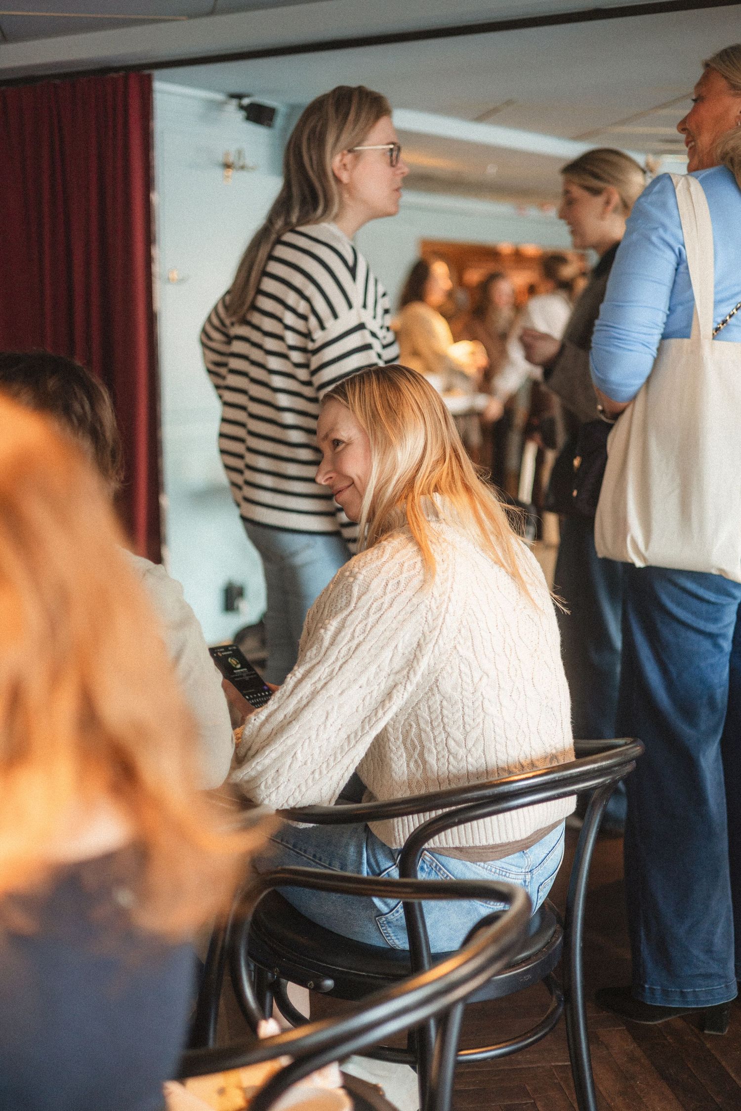 People gather in a casual indoor setting with someone wearing a striped shirt standing and another in a white sweater seated.