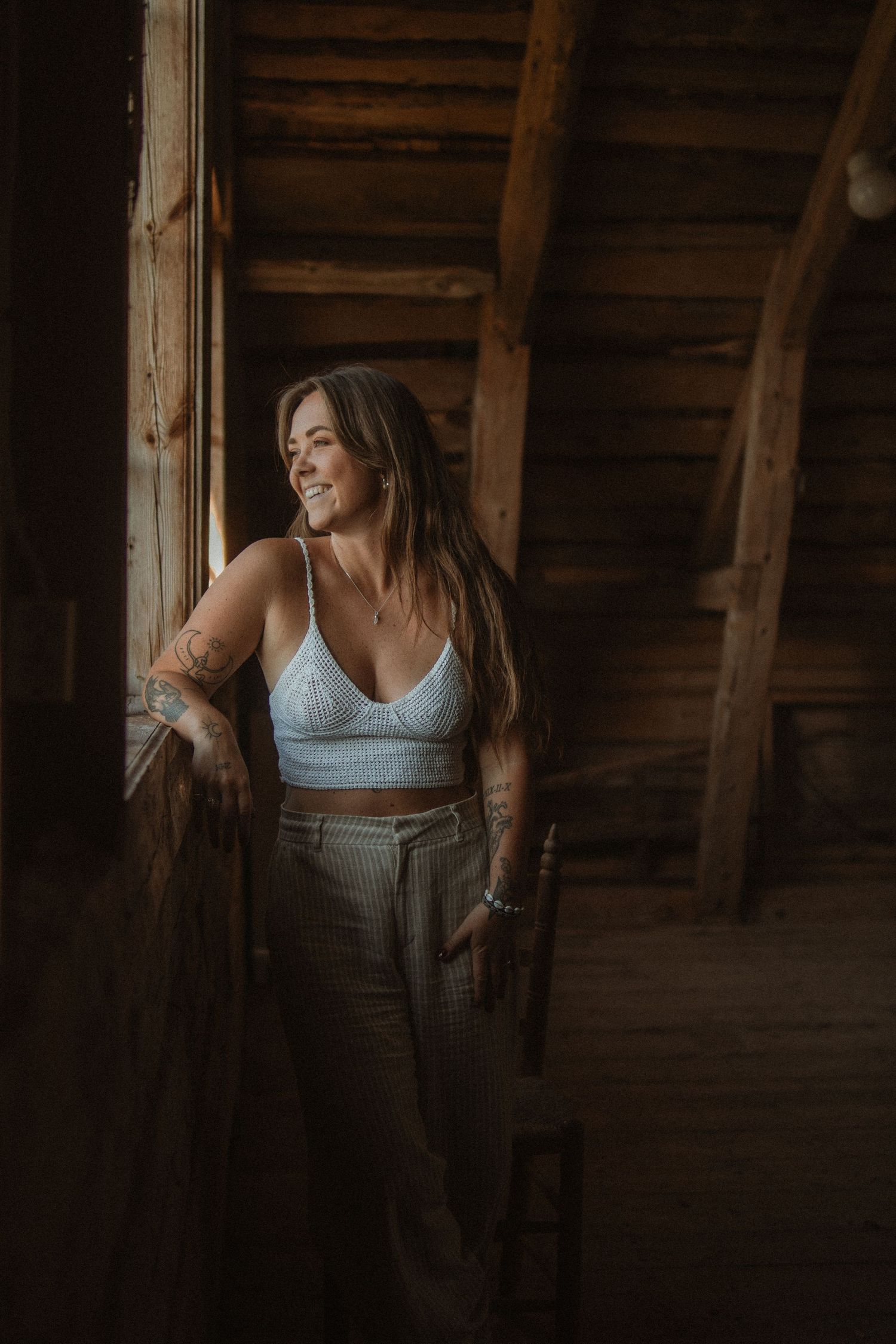 Moody portrait in a rustic wooden attic space featuring a white bralette top and high-waisted pants.