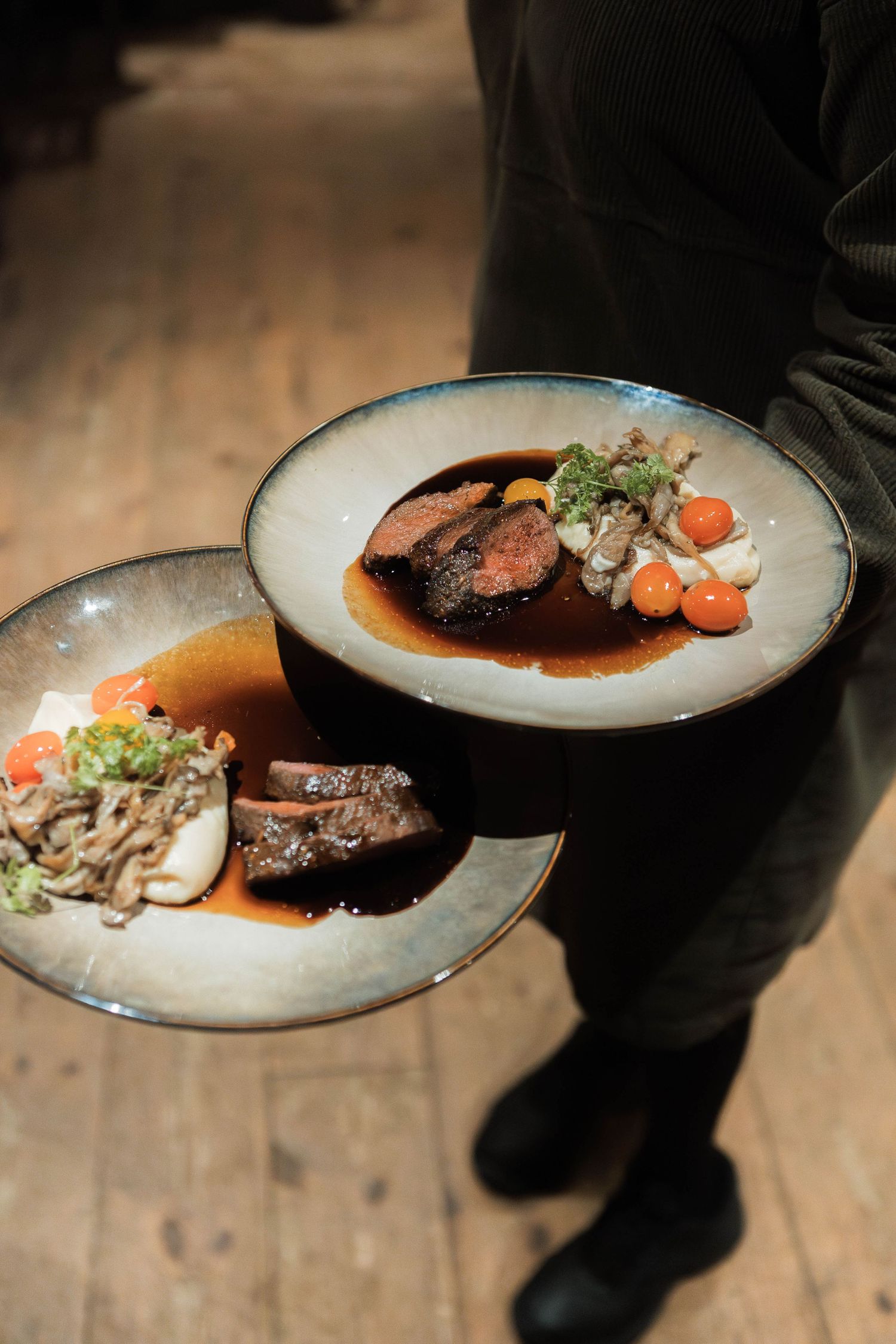 Two elegant plates of gourmet meat dishes with garnishes served on rustic wooden table.
