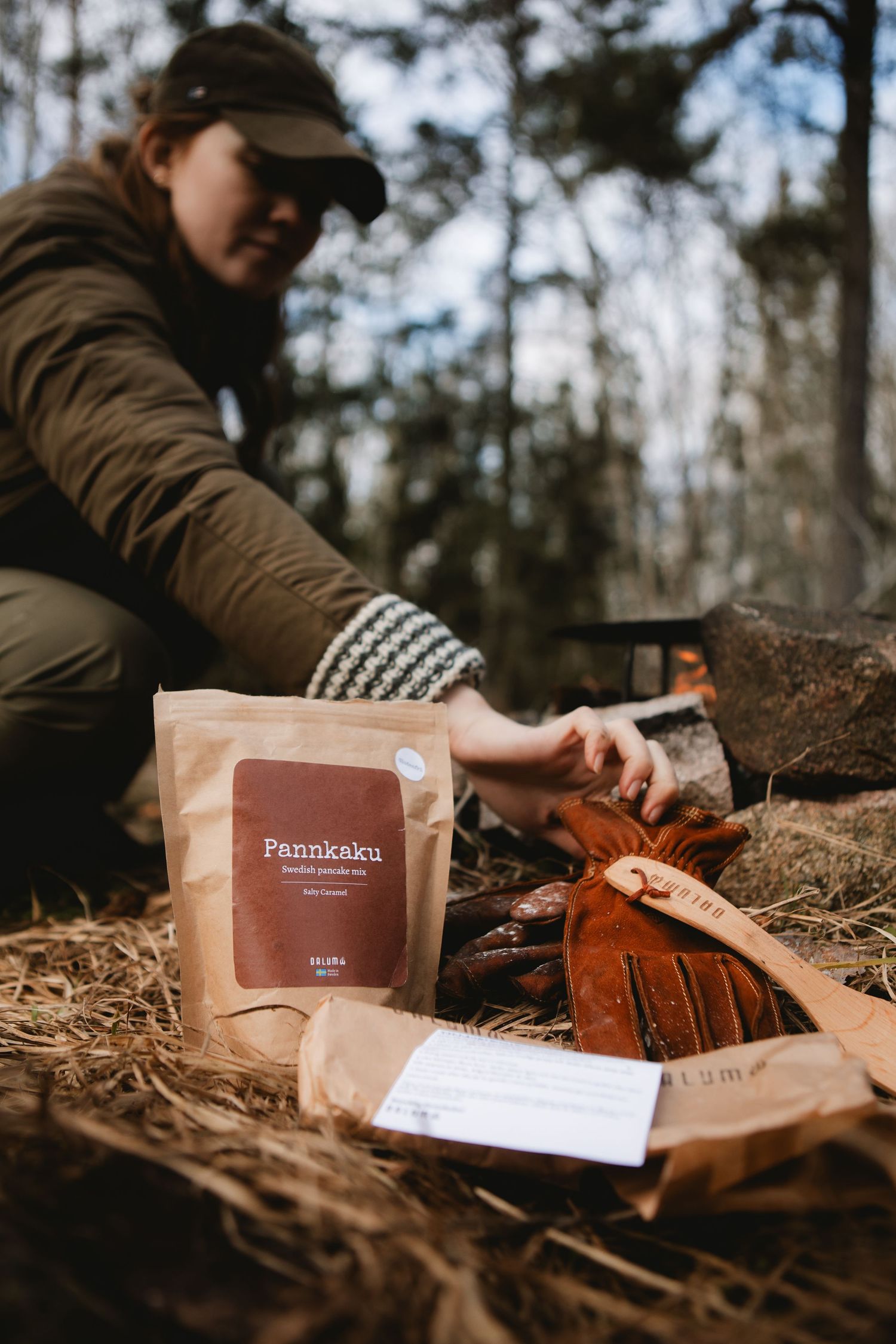 Woman tending a campfire in forest with Pannkaku pancake mix bag and wooden spatula in foreground.