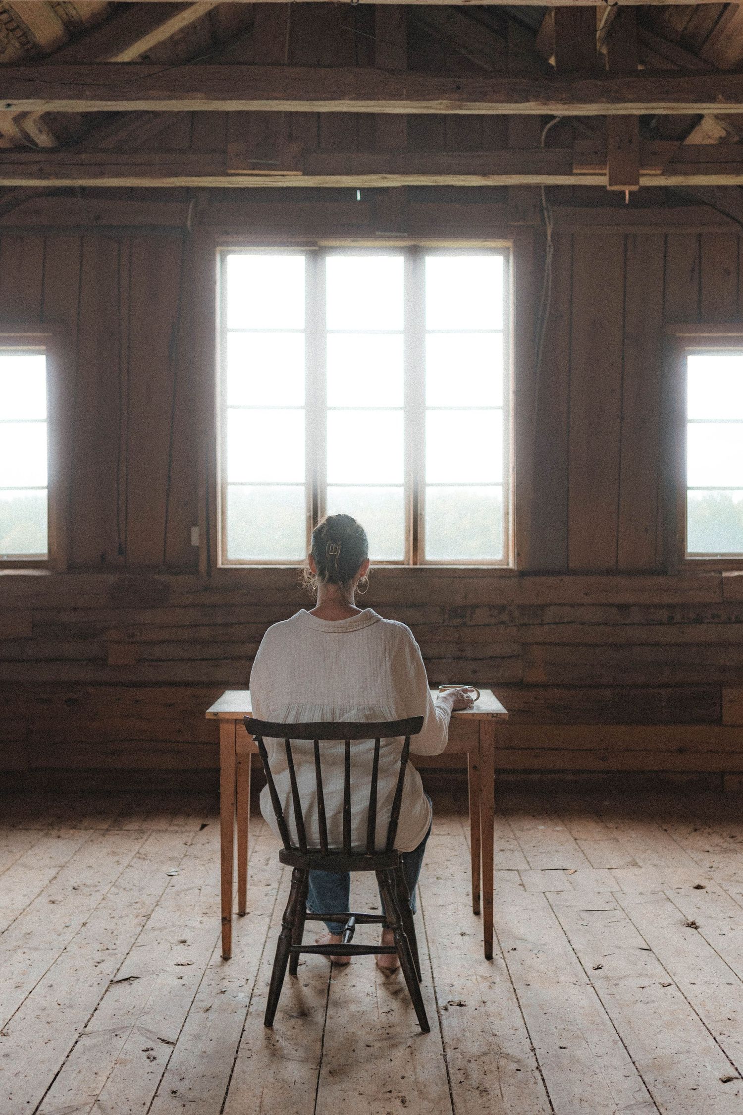 Simple rustic workspace setup with wooden table and black chair near bright windows.