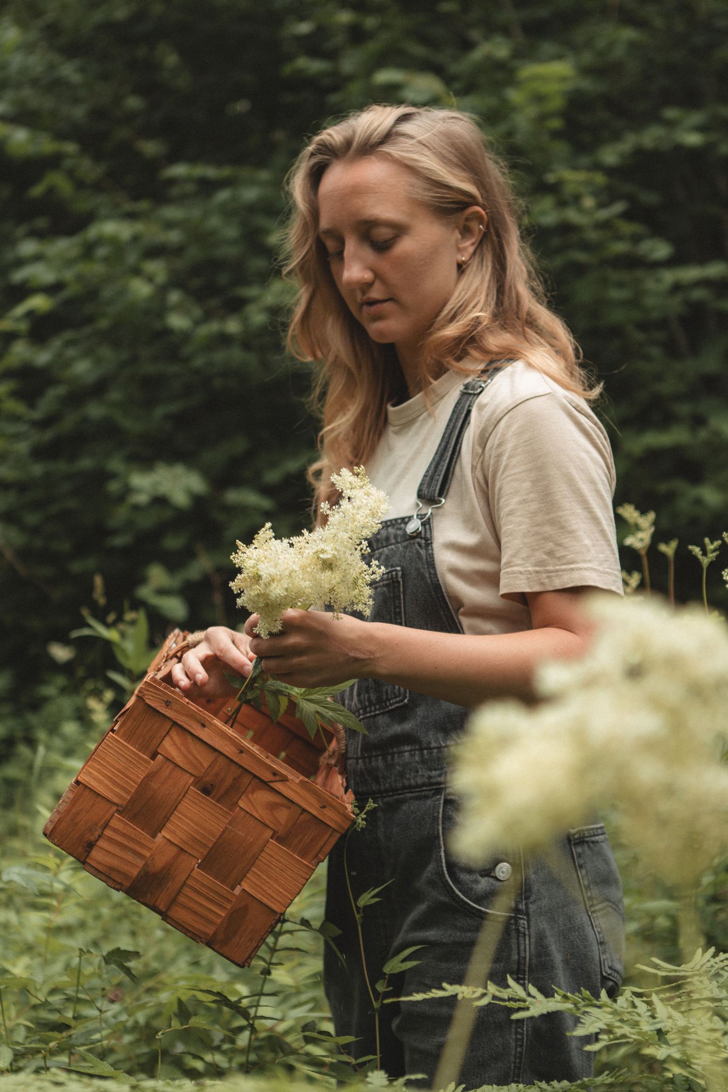 Figure in overalls holding woven basket and wildflowers in green forest setting.
