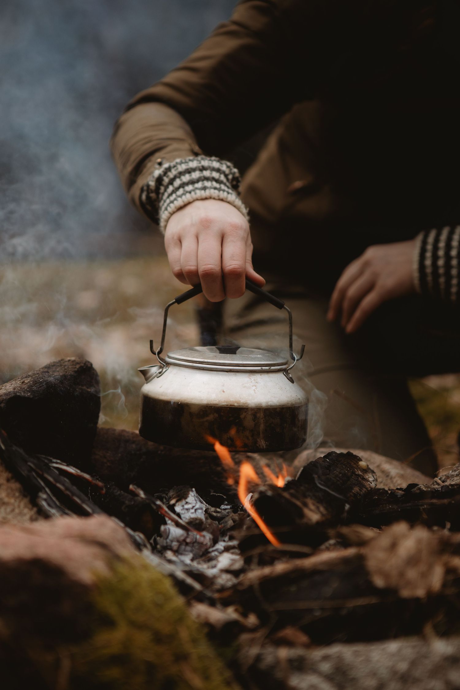 A person wearing bracelets holds a vintage metal kettle over a crackling campfire outdoors.