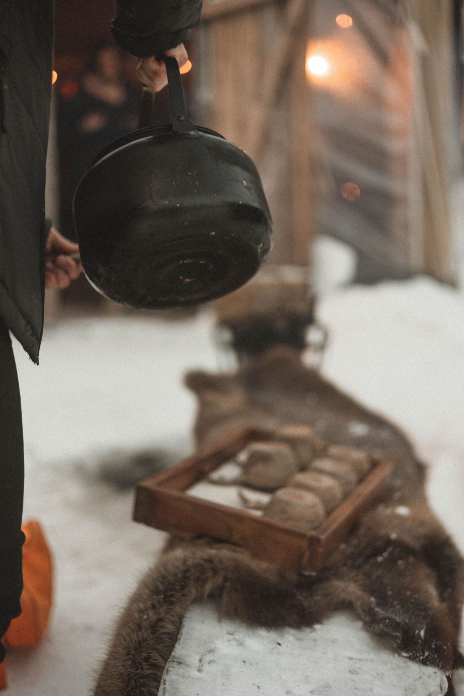 A black cast iron kettle hangs above a rustic wooden display case with tea items in a cozy winter setting.