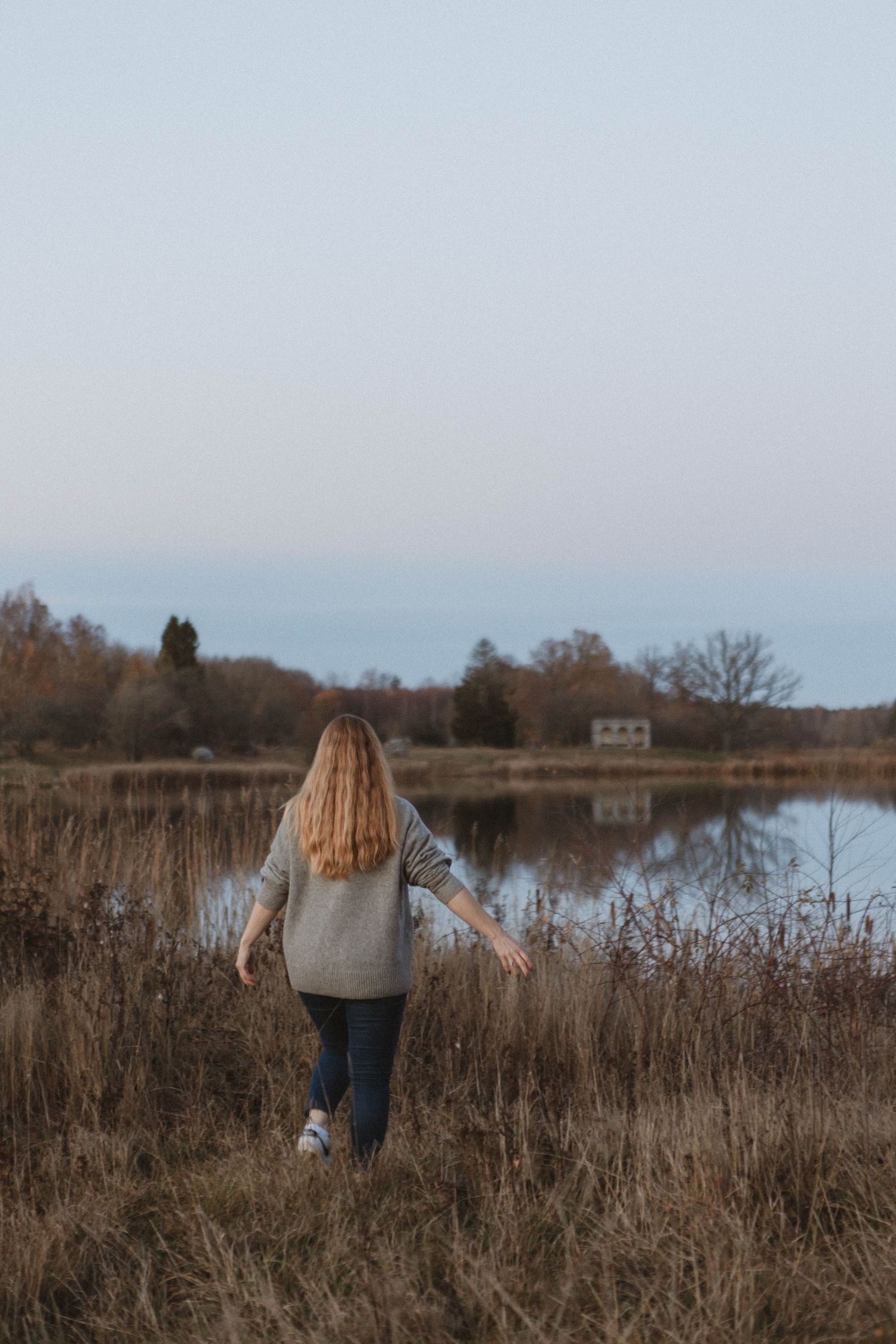 Figure in grey sweater stands near tranquil lake with autumn reeds and trees in background.