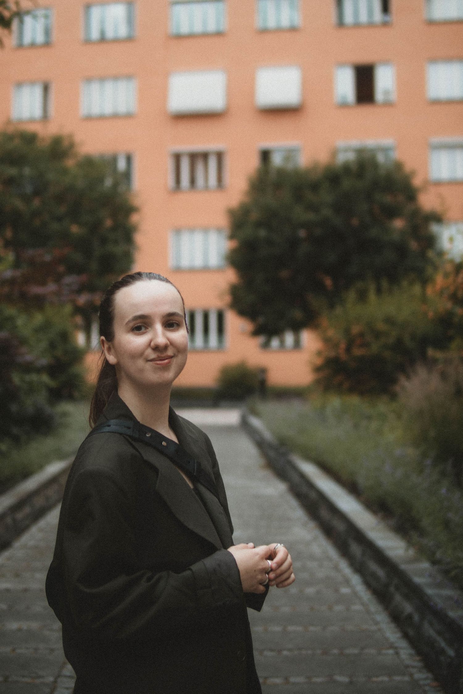 A woman in a black coat stands on a pathway near a peach-colored apartment building with trees in the background.
