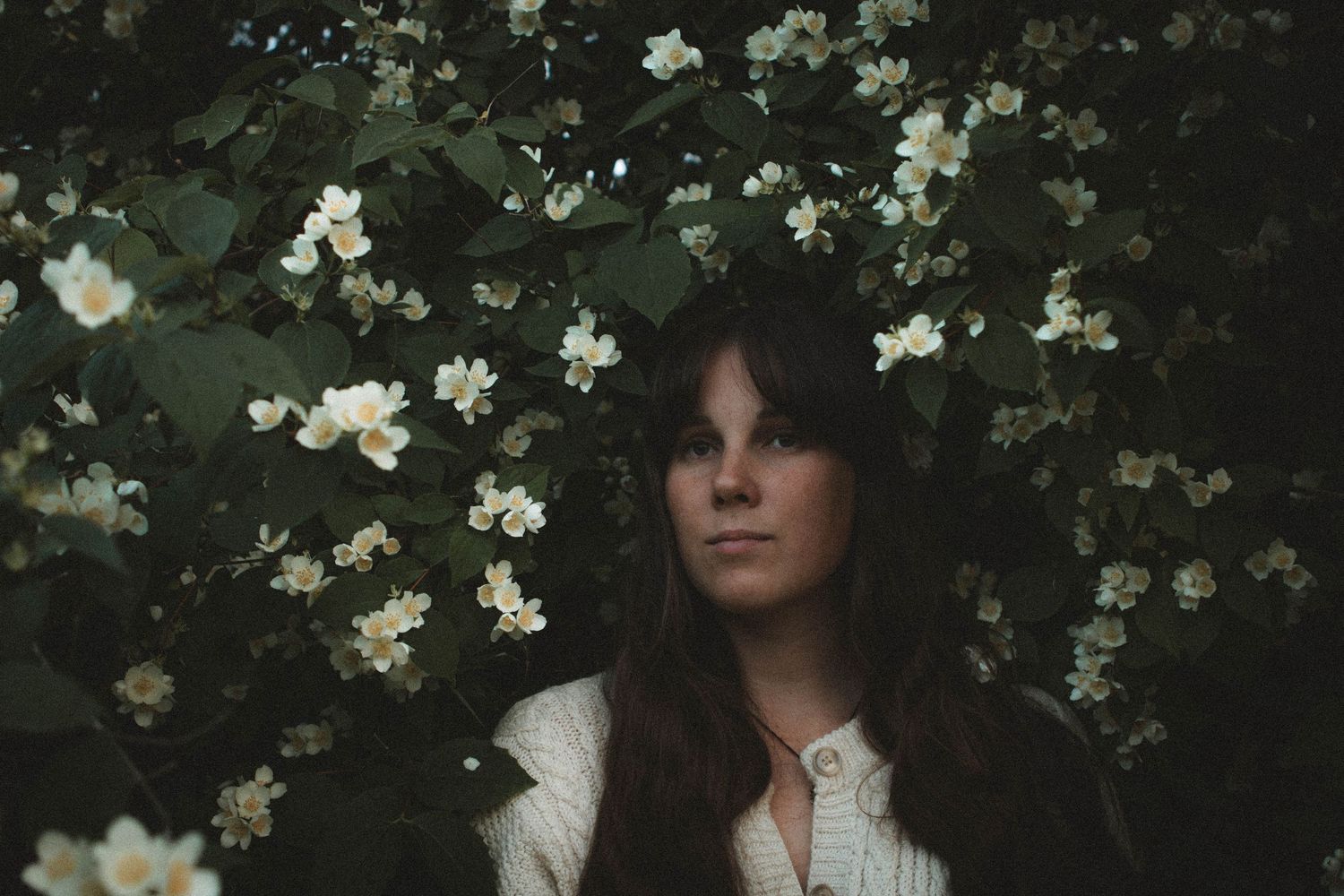 Young woman with long dark hair surrounded by blooming white flowers and lush green leaves in a moody outdoor setting.