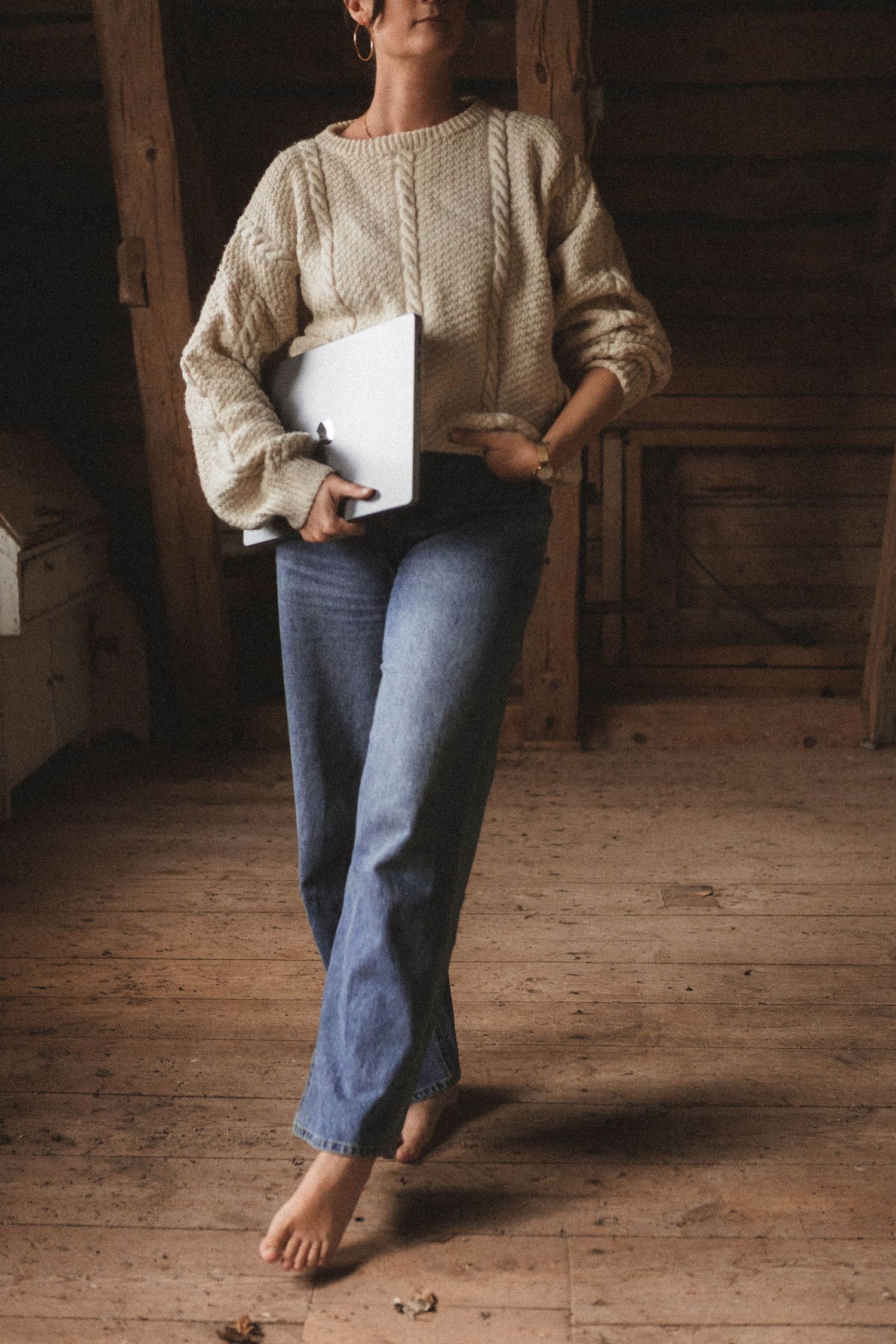 A casual figure in jeans and cream sweater stands barefoot on rustic wooden floorboards.