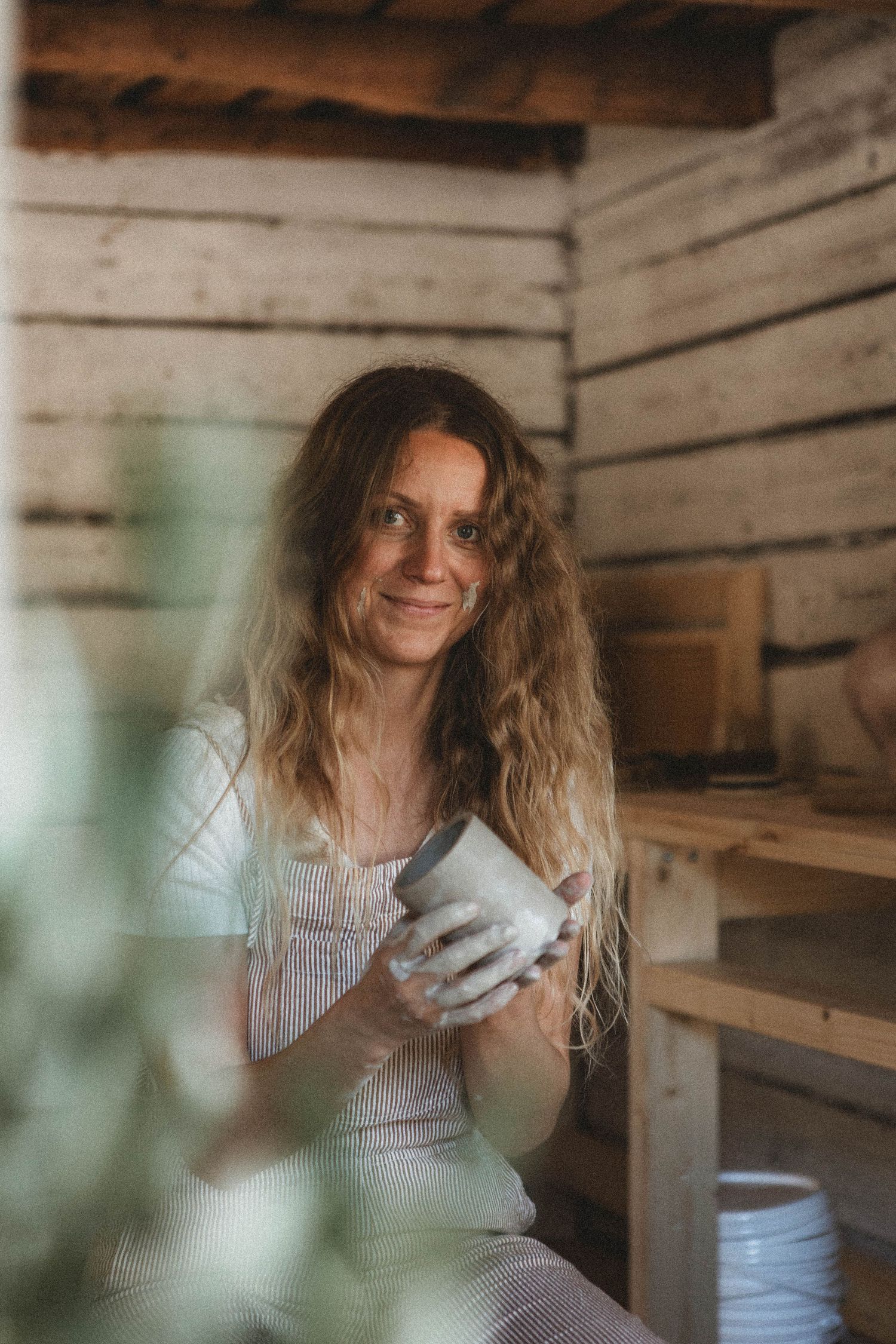 A pottery artist in striped overalls works with clay in a rustic wooden studio, surrounded by shelves of ceramics.