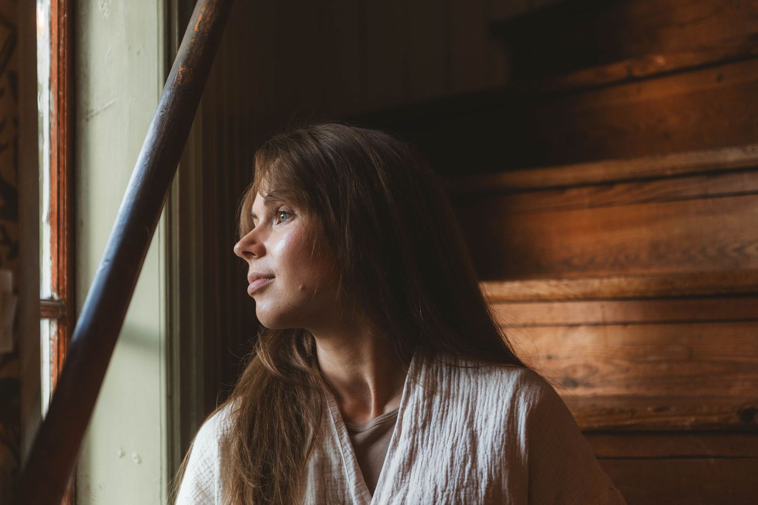Woman in white sweater gazes thoughtfully through window in warm, rustic wooden interior.