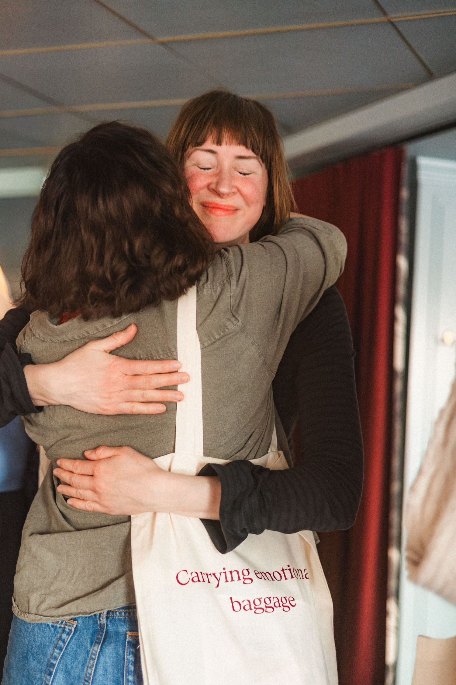 Two people share a warm embrace while one carries a tote bag in an indoor setting.