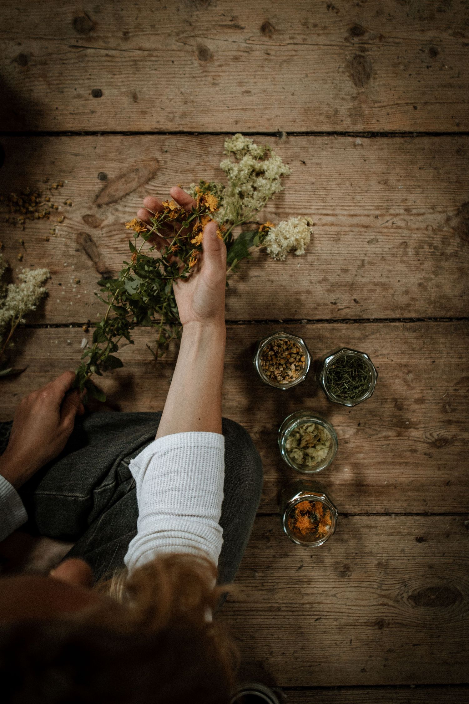 Overhead view of hands arranging flowers on wooden surface with small glass jars.