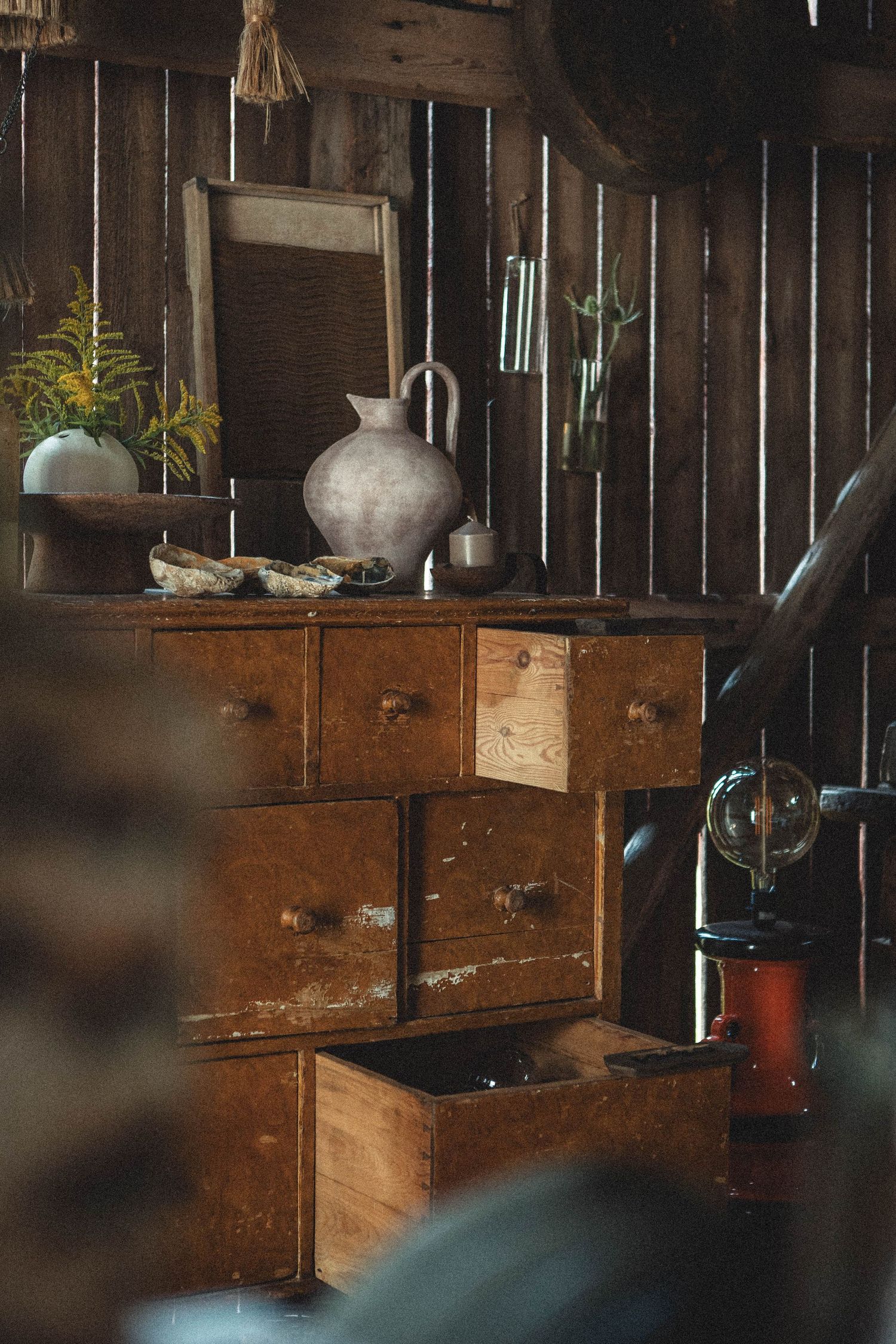 Antique wooden cabinet with drawers displaying ceramic vases and potted plants against wooden wall.