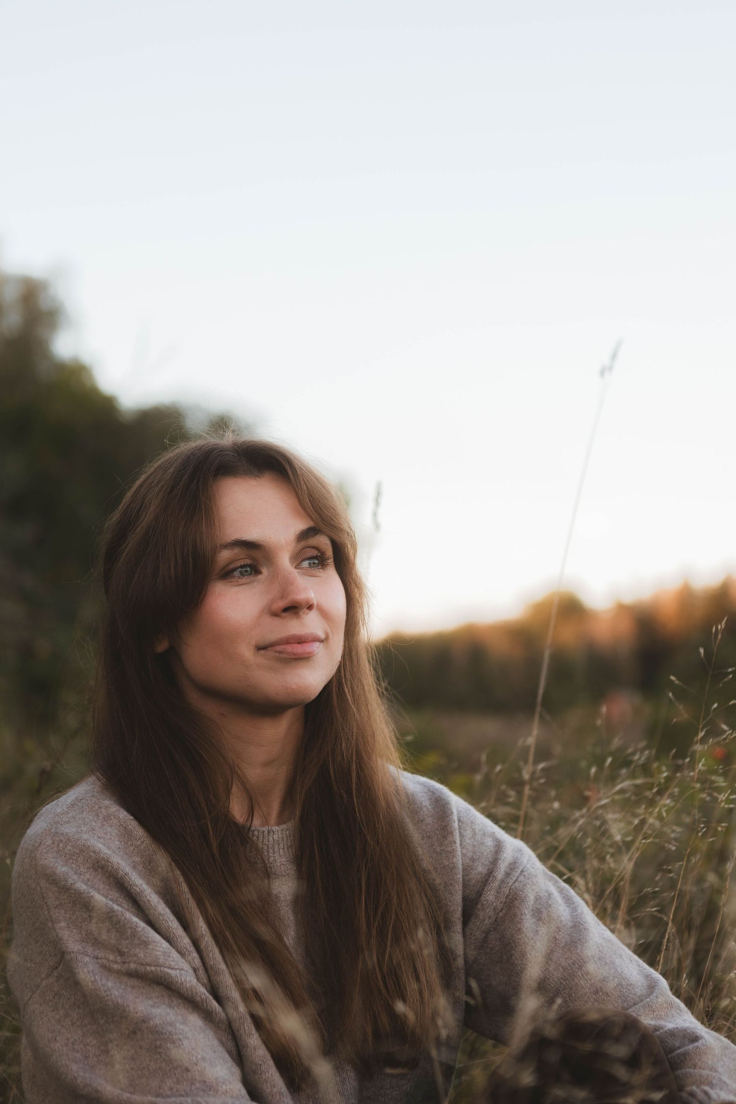 Person smiling contentedly while relaxing in tall grass during sunset wearing gray sweater.