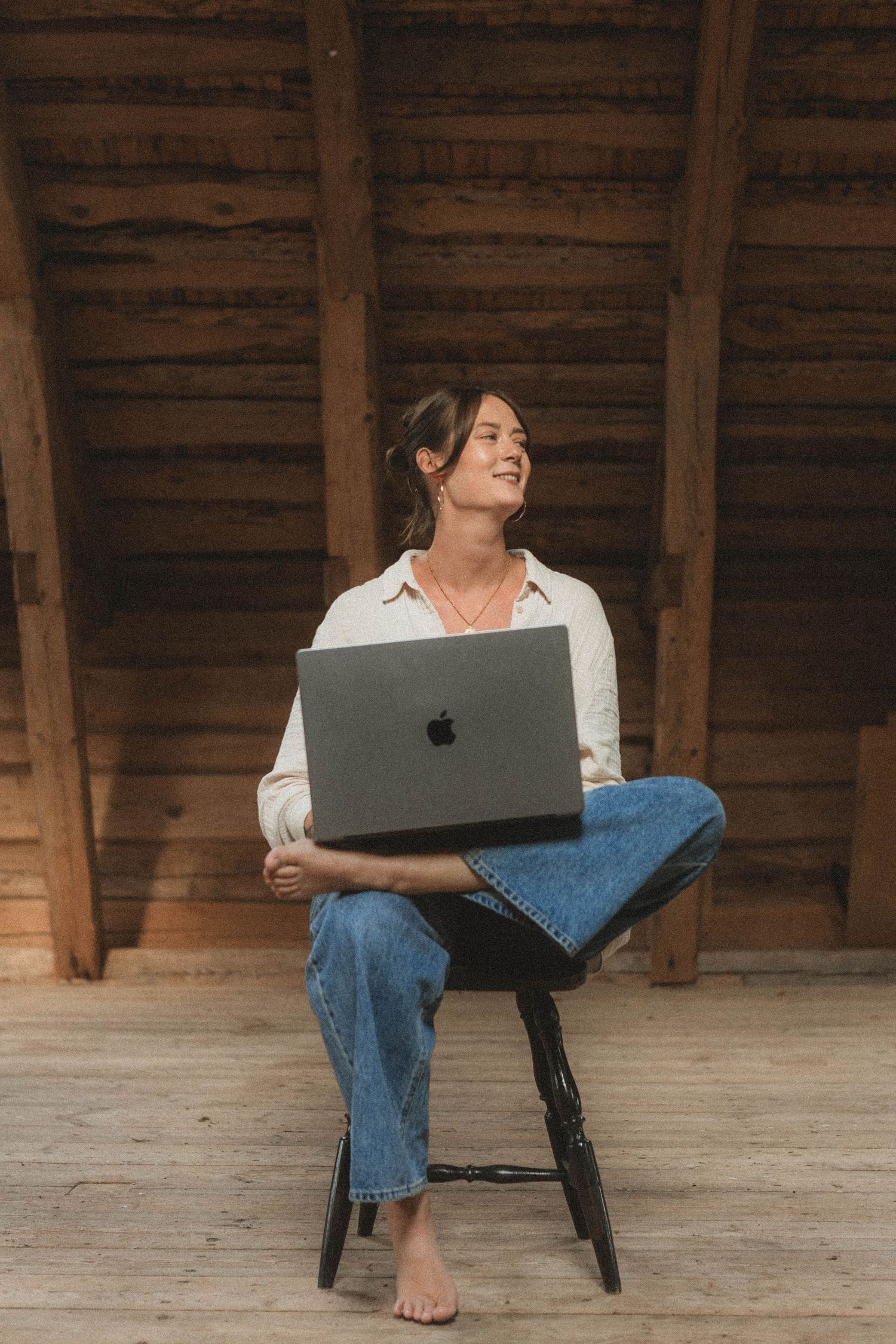 Person wearing white shirt and jeans works casually on laptop while sitting on wooden floor in rustic space.