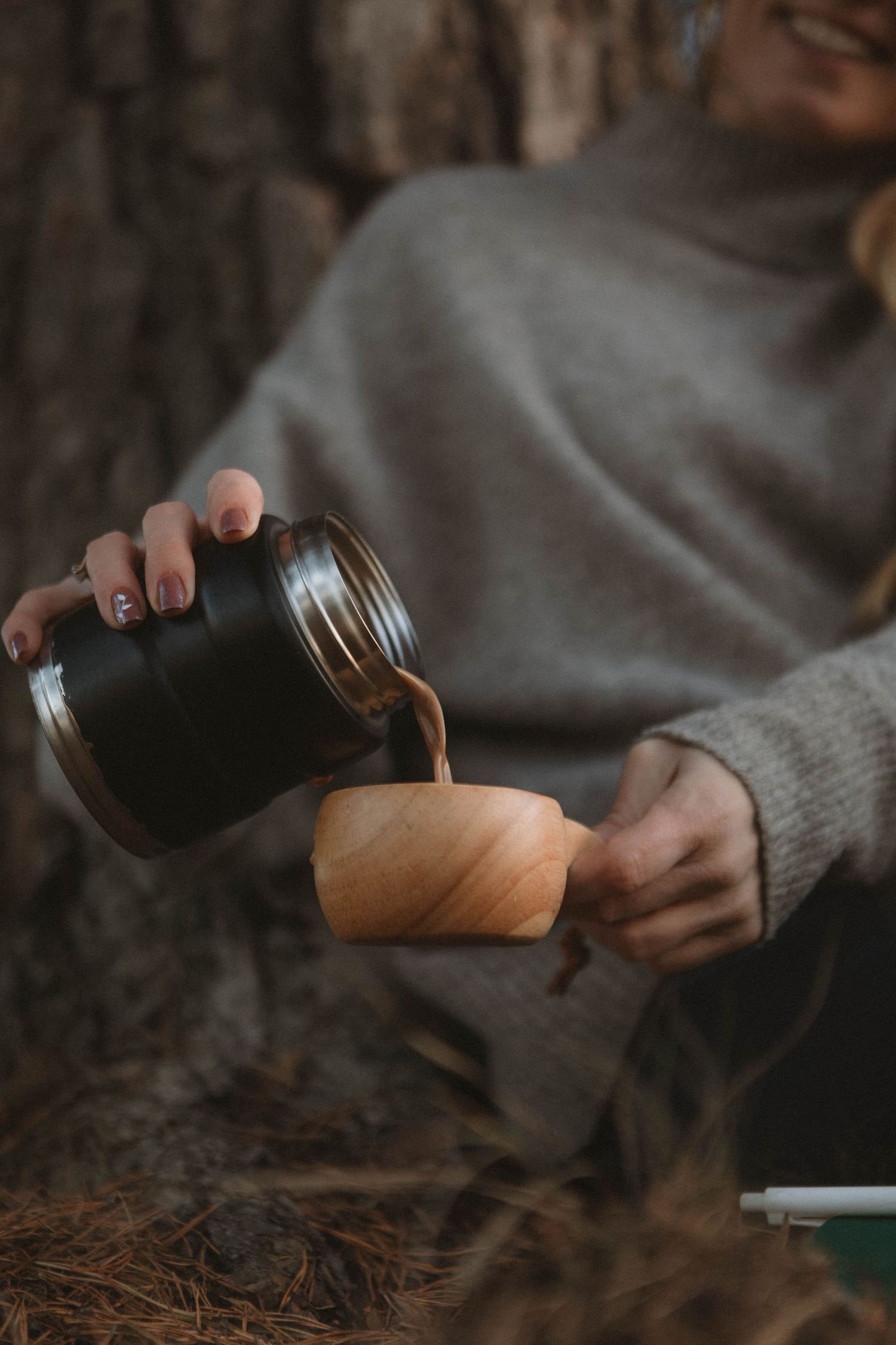 Someone in a grey sweater pours a hot beverage into a wooden cup in a cozy rustic setting.