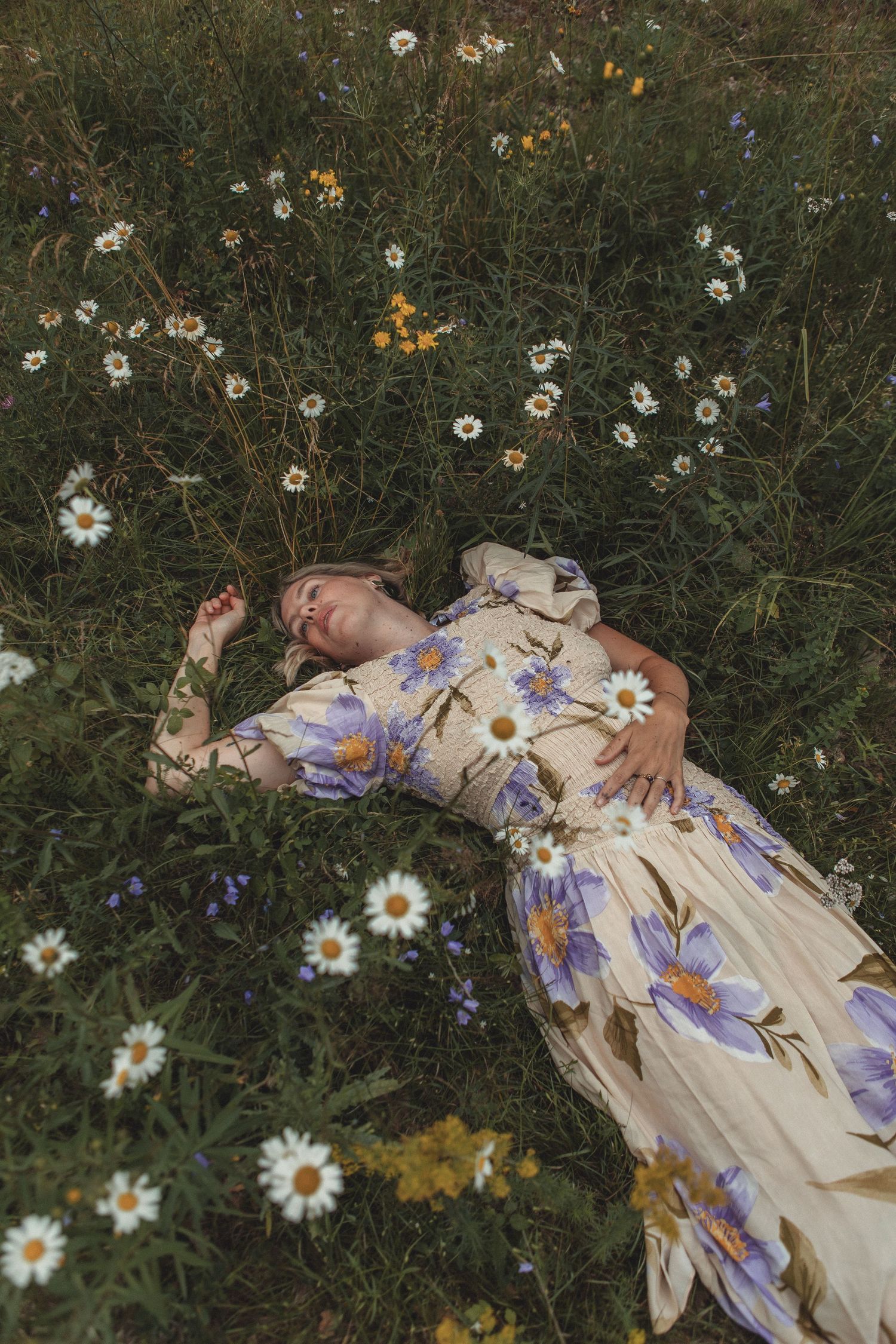 Someone lies in a meadow surrounded by daisies wearing a floral dress during summer.