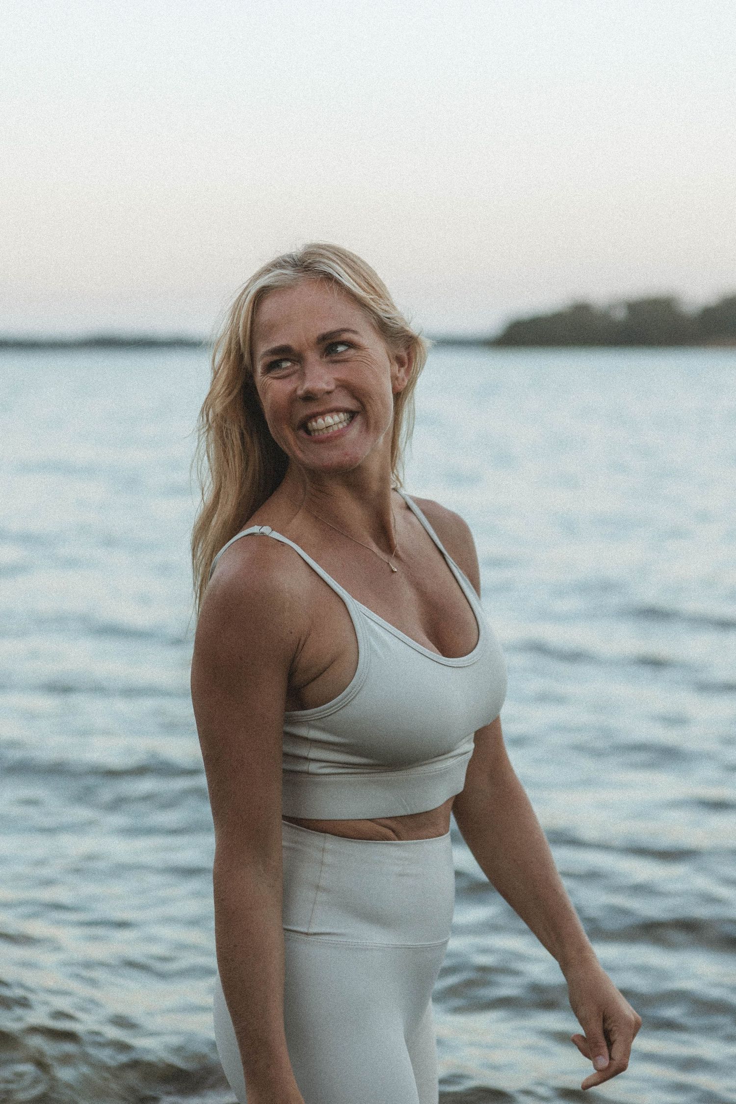 Person in white workout set smiles while enjoying coastal sunset.