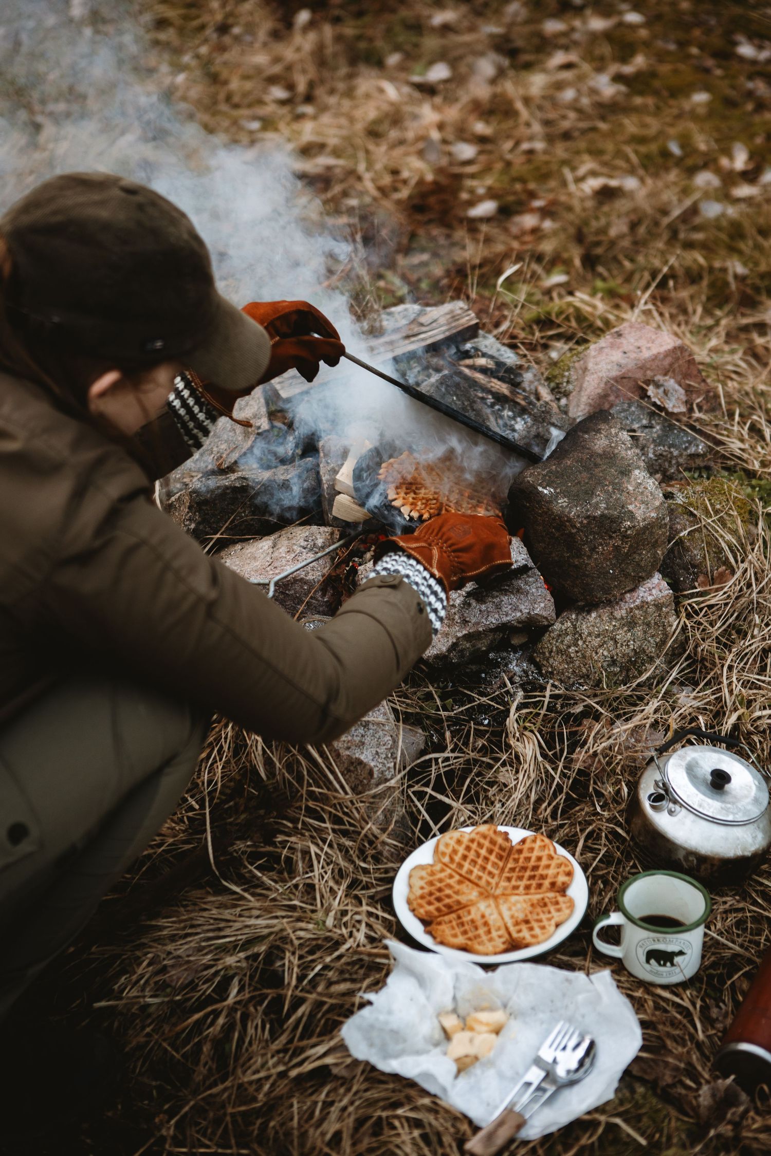 Person cooking over a campfire with waffles, coffee, and utensils on the ground nearby.