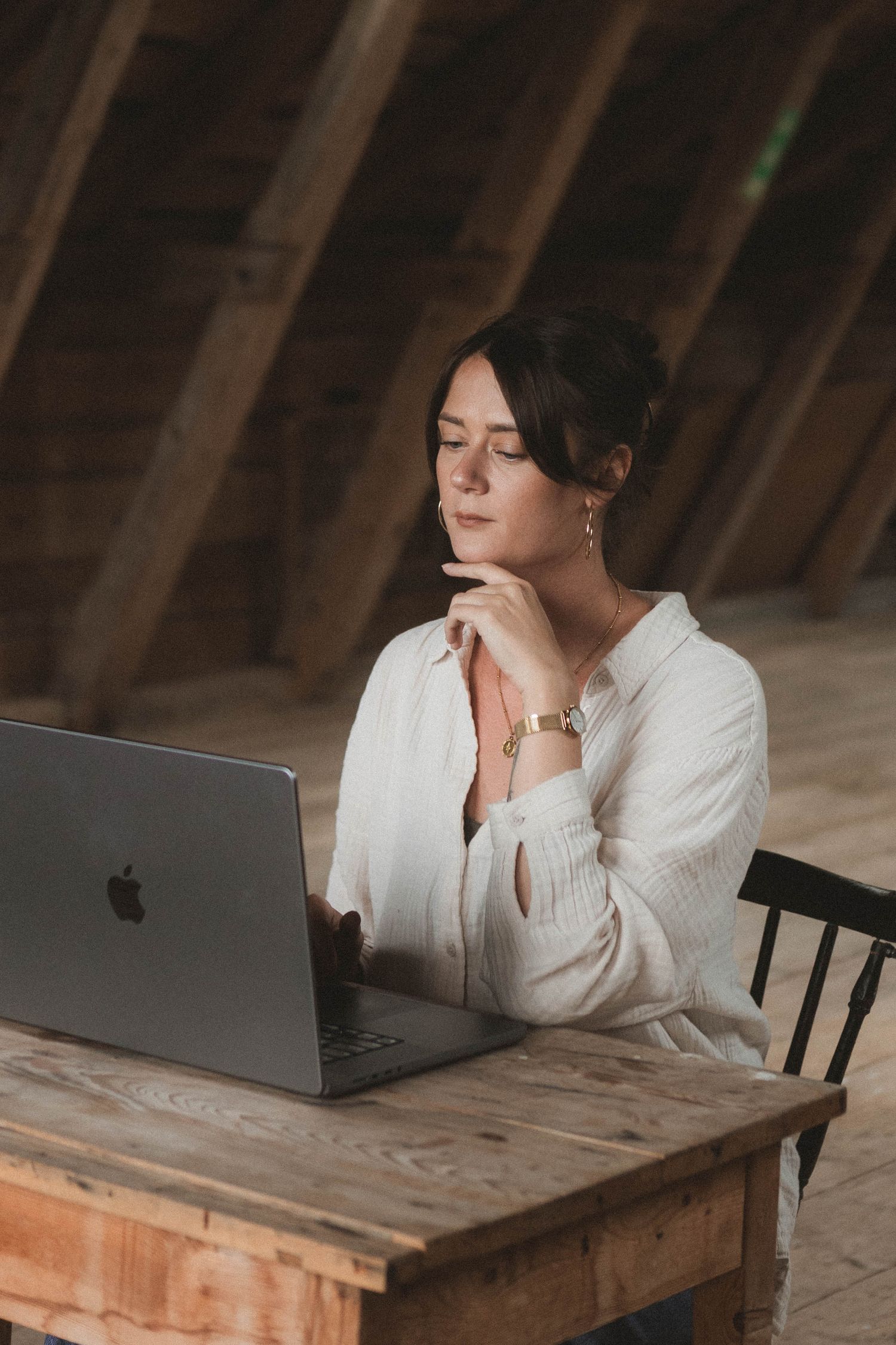 Person working on MacBook laptop at wooden desk in atmospheric attic setting.
