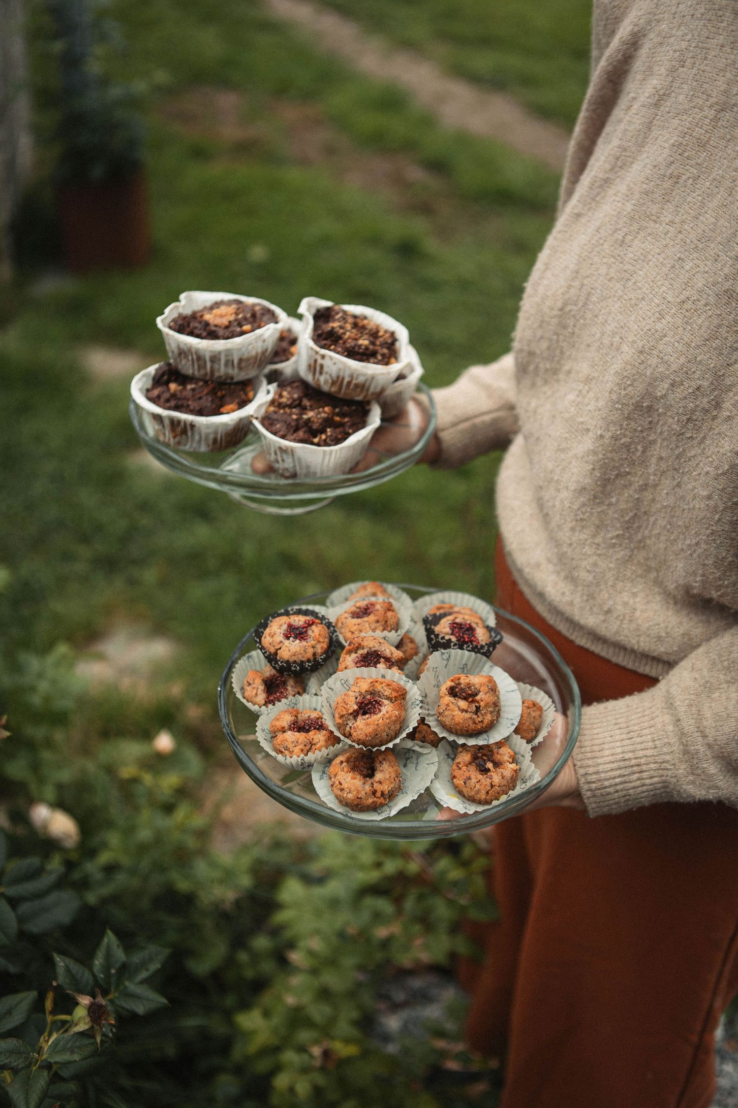 Two-tiered serving platter displaying homemade muffins and donuts in paper liners outdoors.