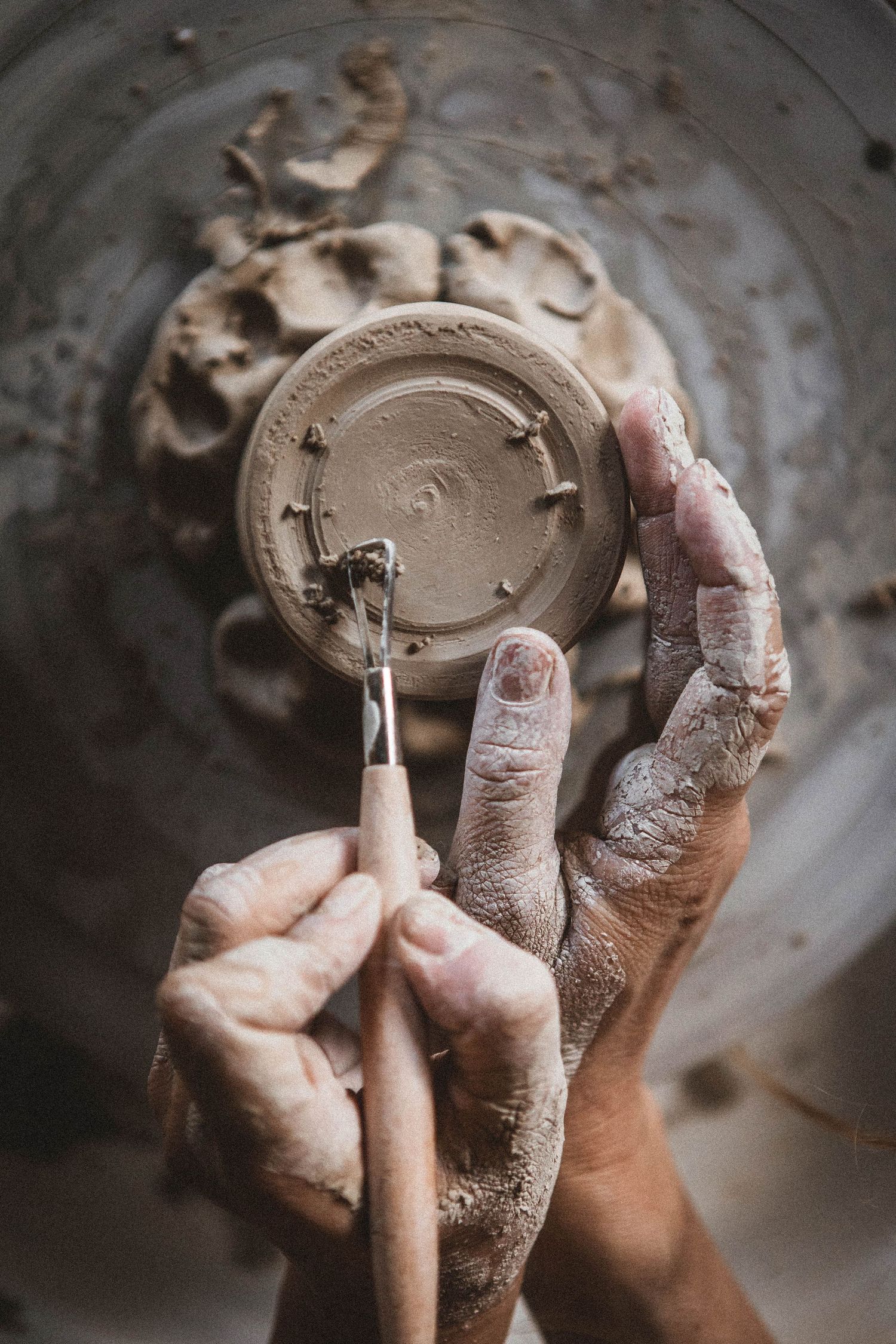 Close-up of hands carving details into base of clay vessel with pottery tools.