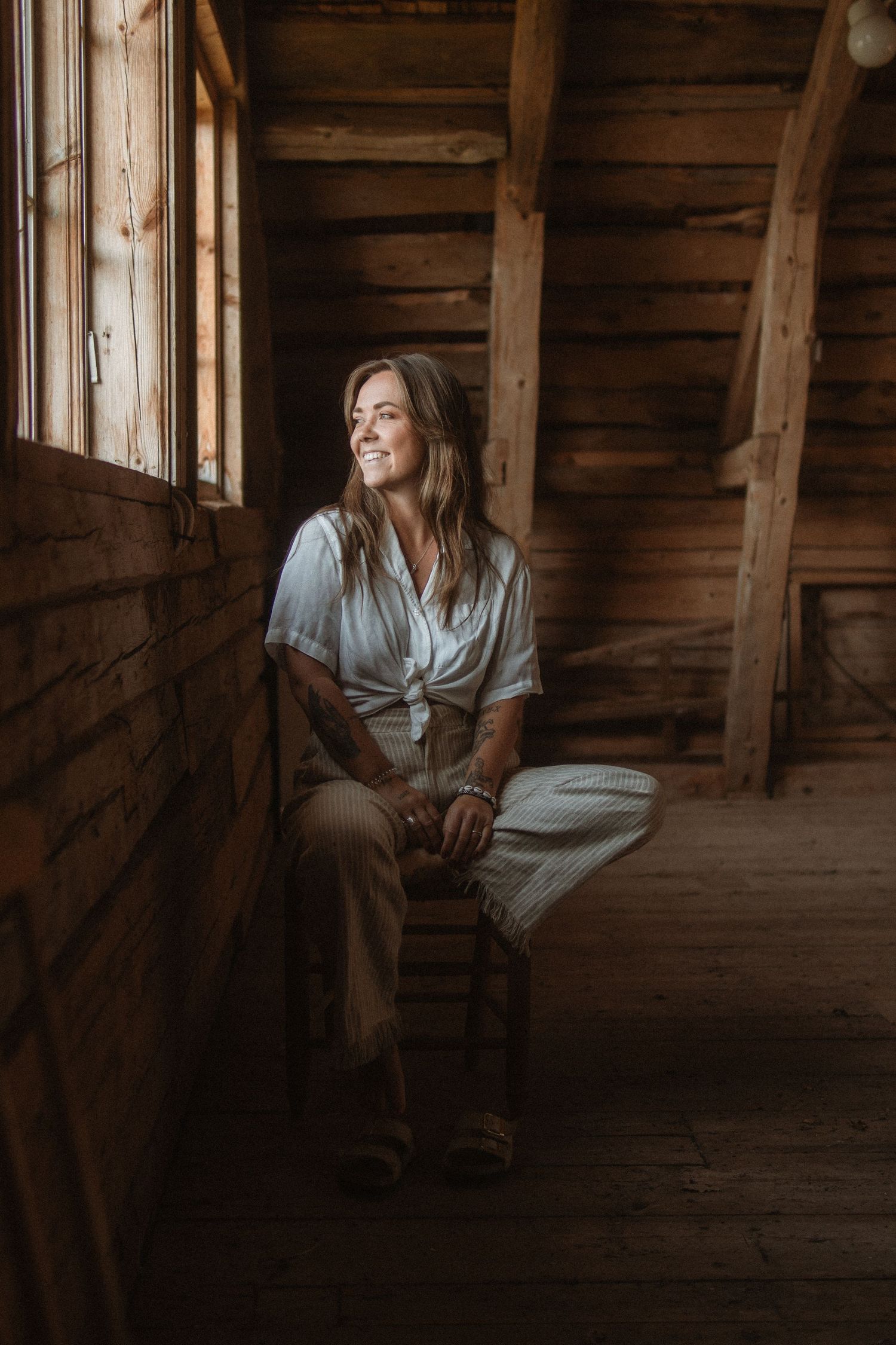 A figure sits by a rustic wooden window in a barn, wearing a light colored top and looking toward natural light.