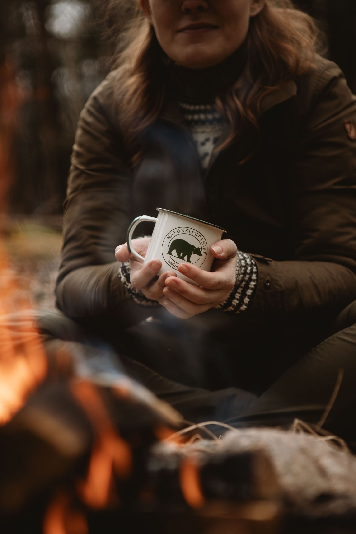 Woman holding a steaming enamel bear mug by a campfire in a forest, wearing a cozy jacket and beaded bracelet.