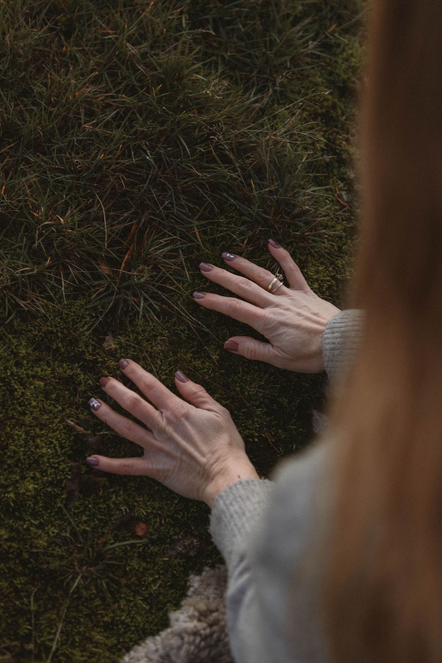 Two hands wearing a beige sweater reach out to touch a mossy green forest floor in a moody nature photograph.